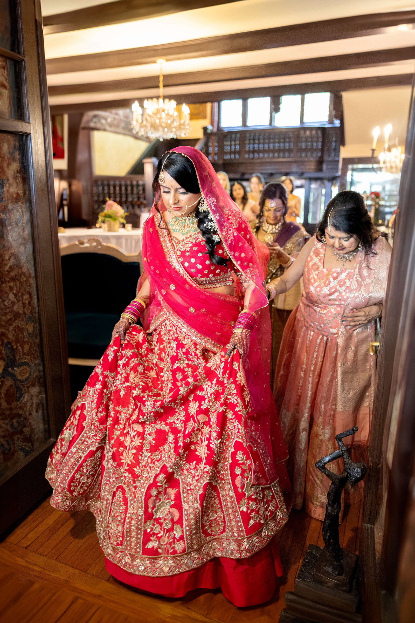 A bride in a vibrant red and gold embroidered lehenga is being assisted by two women in a richly decorated room with chandeliers and wood-paneled walls. She wears traditional jewelry, and the scene conveys a sense of preparation and celebration.