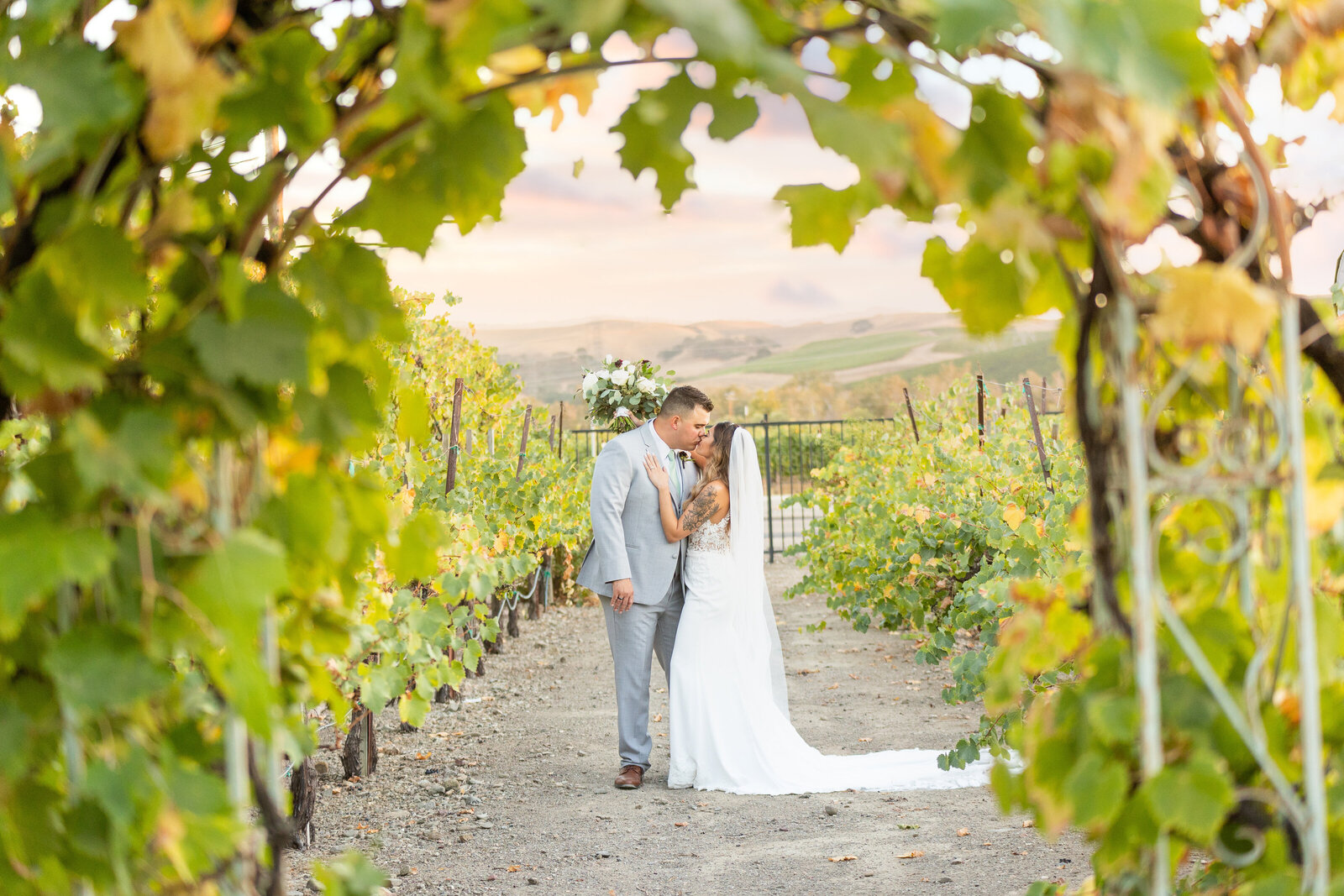 Bride and groom kiss in the middle of a vineyard with a leaf arch in the foreground, photo by wedding photographer sacramento, ca, philippe studio pro.