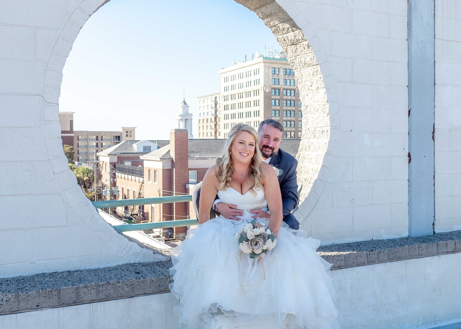 The bride and groom pose together on a rooftop in downtown Wilmington, NC, framed by a circular architectural feature with city buildings in the background, blending urban charm and romantic elegance.