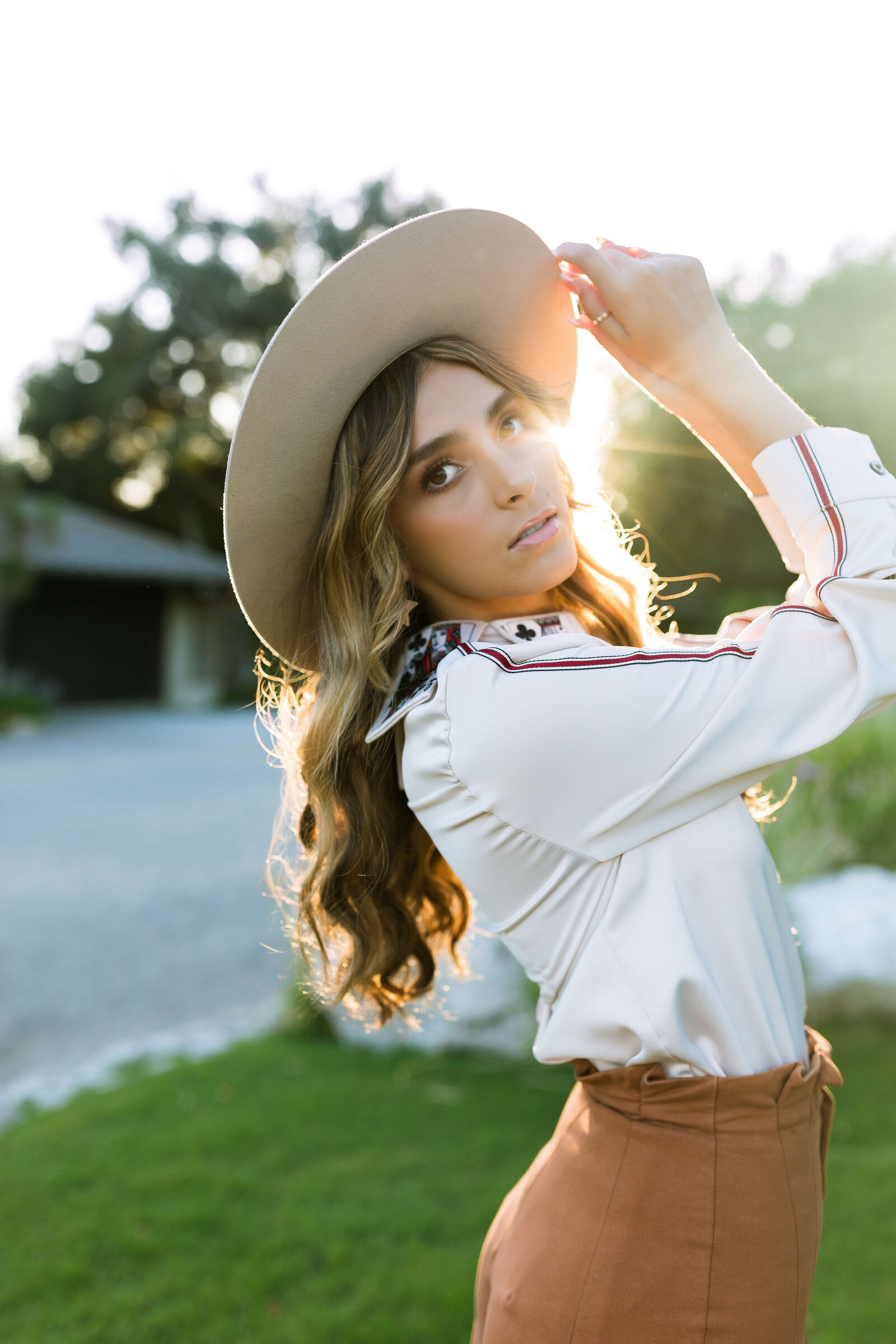 brunette girl with highlights wearing white top and brown bottoms and holding her hat