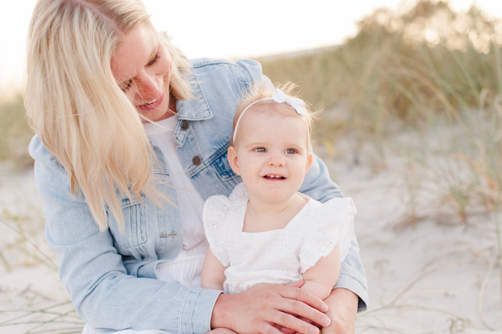 Blonde mom sits in the dunes holding her young daughter and smiling down at her during golden hour