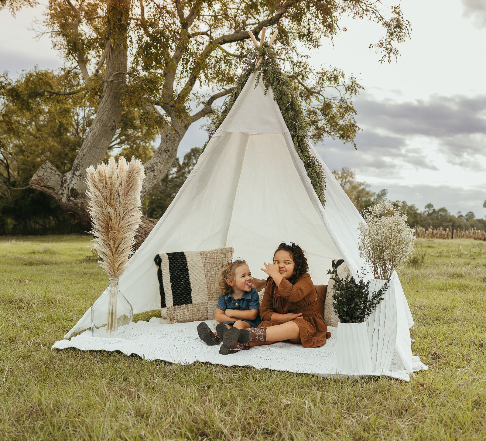 Family session in a farm field in Aiken SC - two siblings sitting together inside a boho white teepee with pampas grass decor.