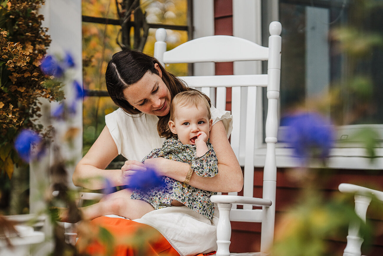 mother sits in white wooden rocking chair on porch with baby surrounded by purple flowers