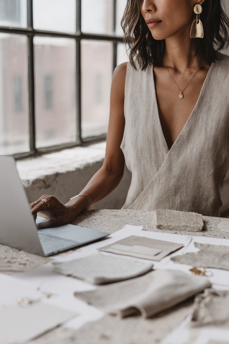 Editorial photograph of a woman designer working on texture samples and a laptop in natural light, capturing the refined, organic design process of Kathalyst Design Studio.
