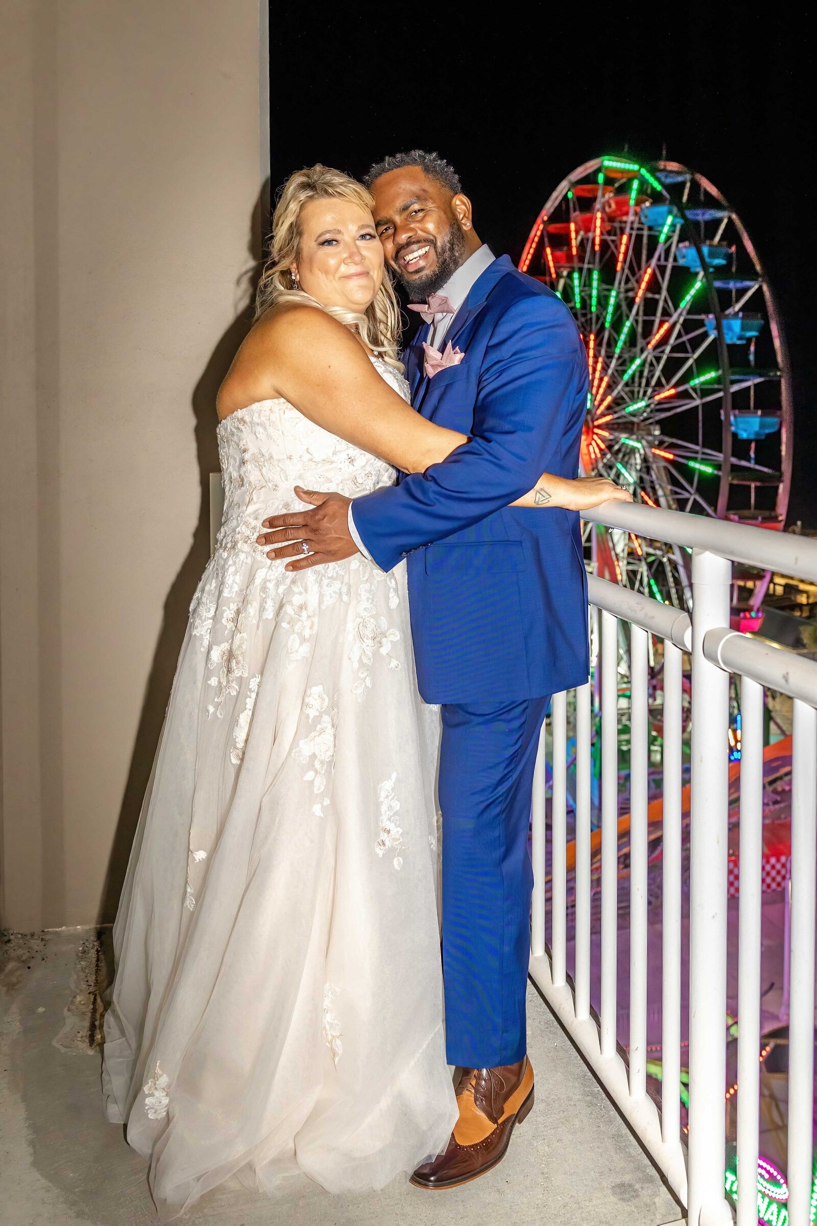 The bride and groom embrace on a balcony overlooking a brightly lit Ferris wheel at night, captured during a Carolina Beach, NC wedding, blending romance with vibrant city charm.