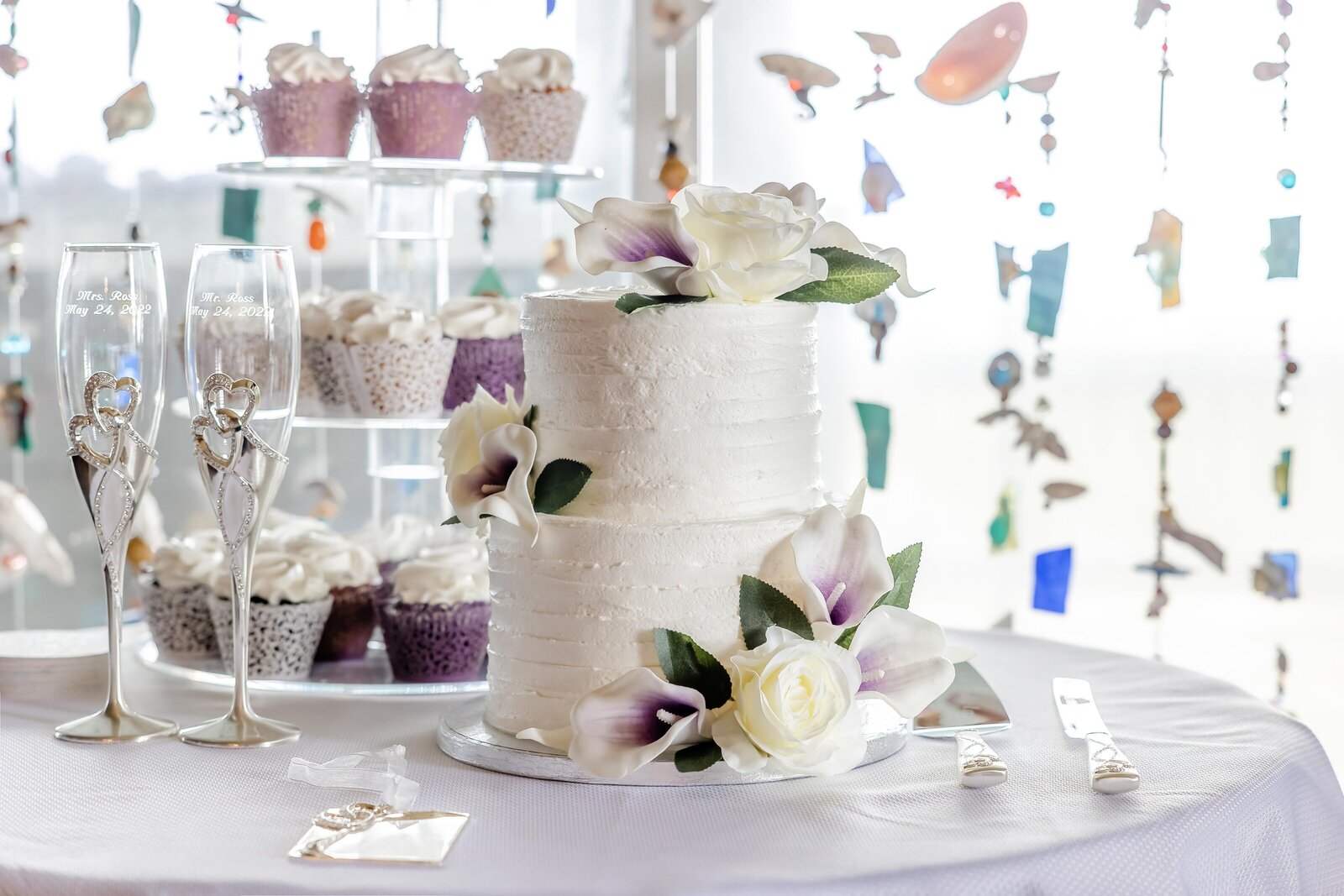 A white tiered wedding cake with purple floral accents sits next to matching cupcakes and champagne flutes, against a backdrop of coastal-inspired hanging decor, captured during an Outer Banks, NC wedding.