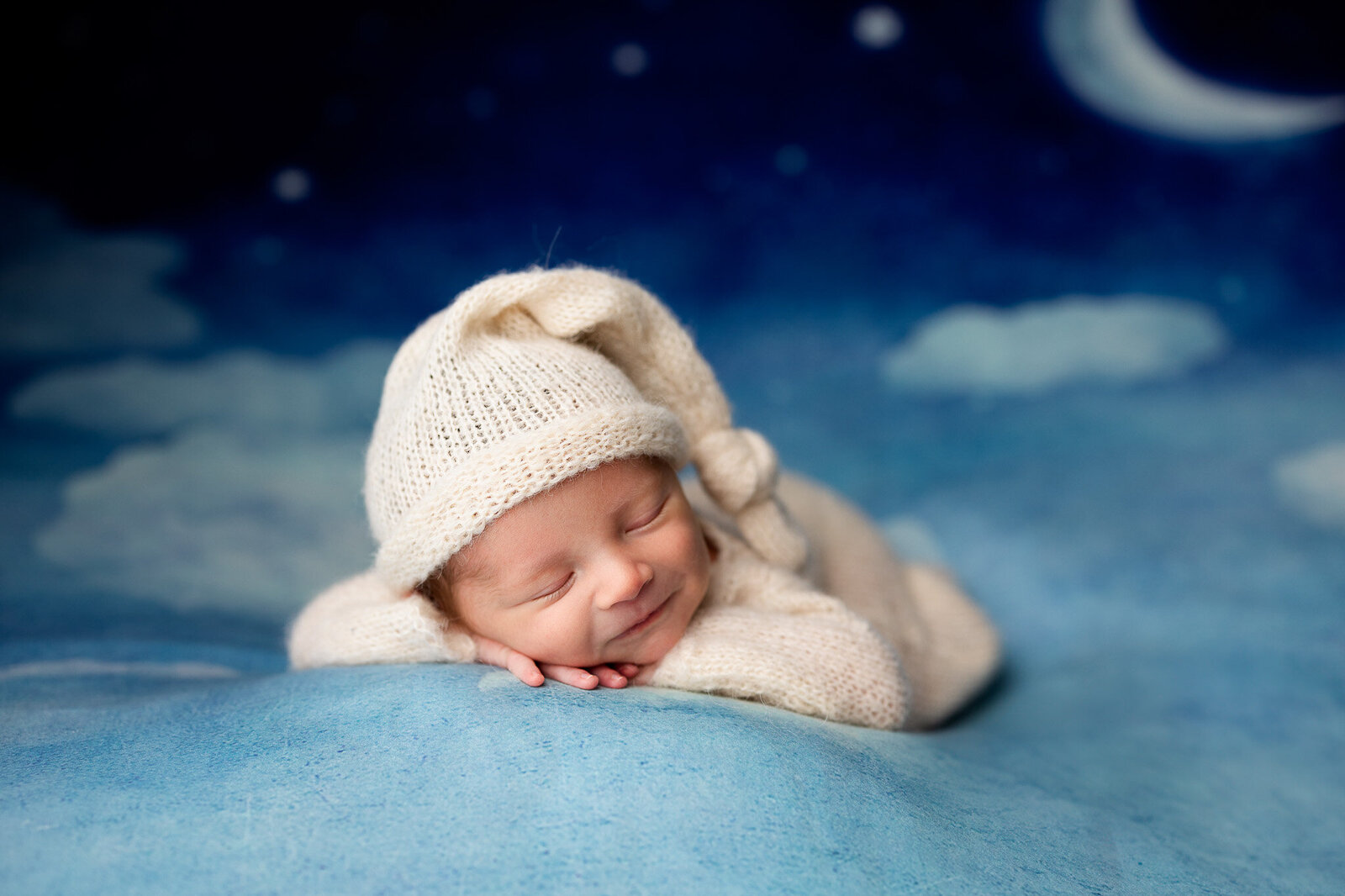 Baby boy in a cream knit sleeper and night cap, posed forward facing with chin on hands in front of a night sky backdrop