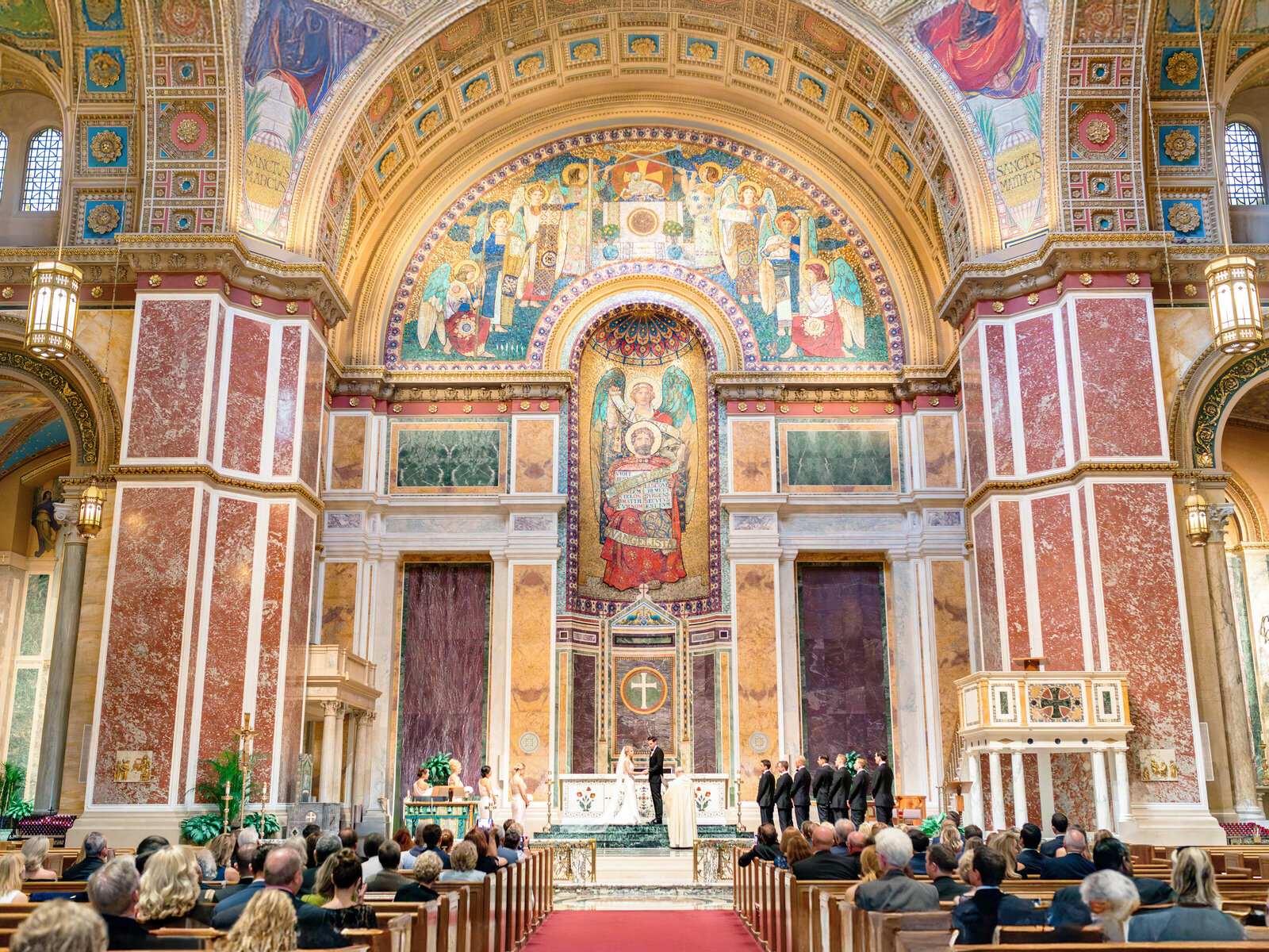 A grand interior of a cathedral with an elaborate domed ceiling featuring religious mosaics. The altar is central, with clergy and choir members present. Seated attendees fill the pews, observing the ceremony. The setting is ornate and classical.