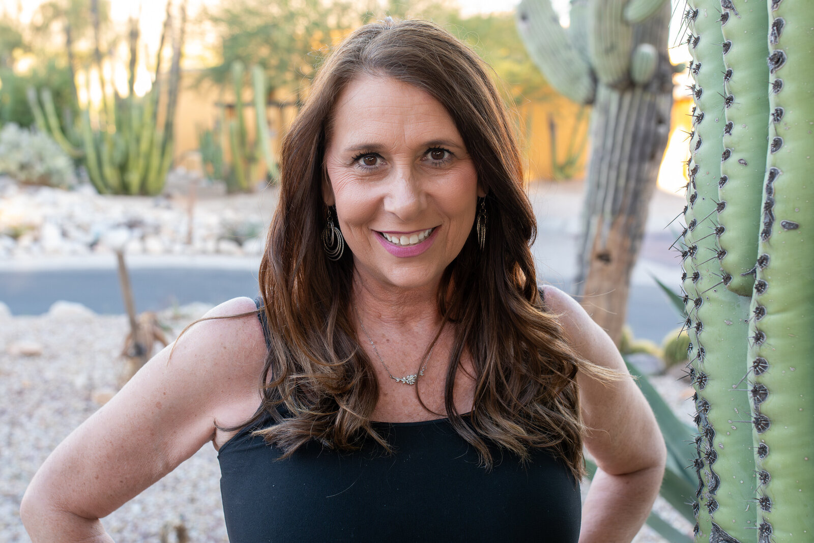Smiling woman with long brown hair wearing a black top beside a tall cactus, photographed by Vyrl Photo, showcasing Tucson brand photography.