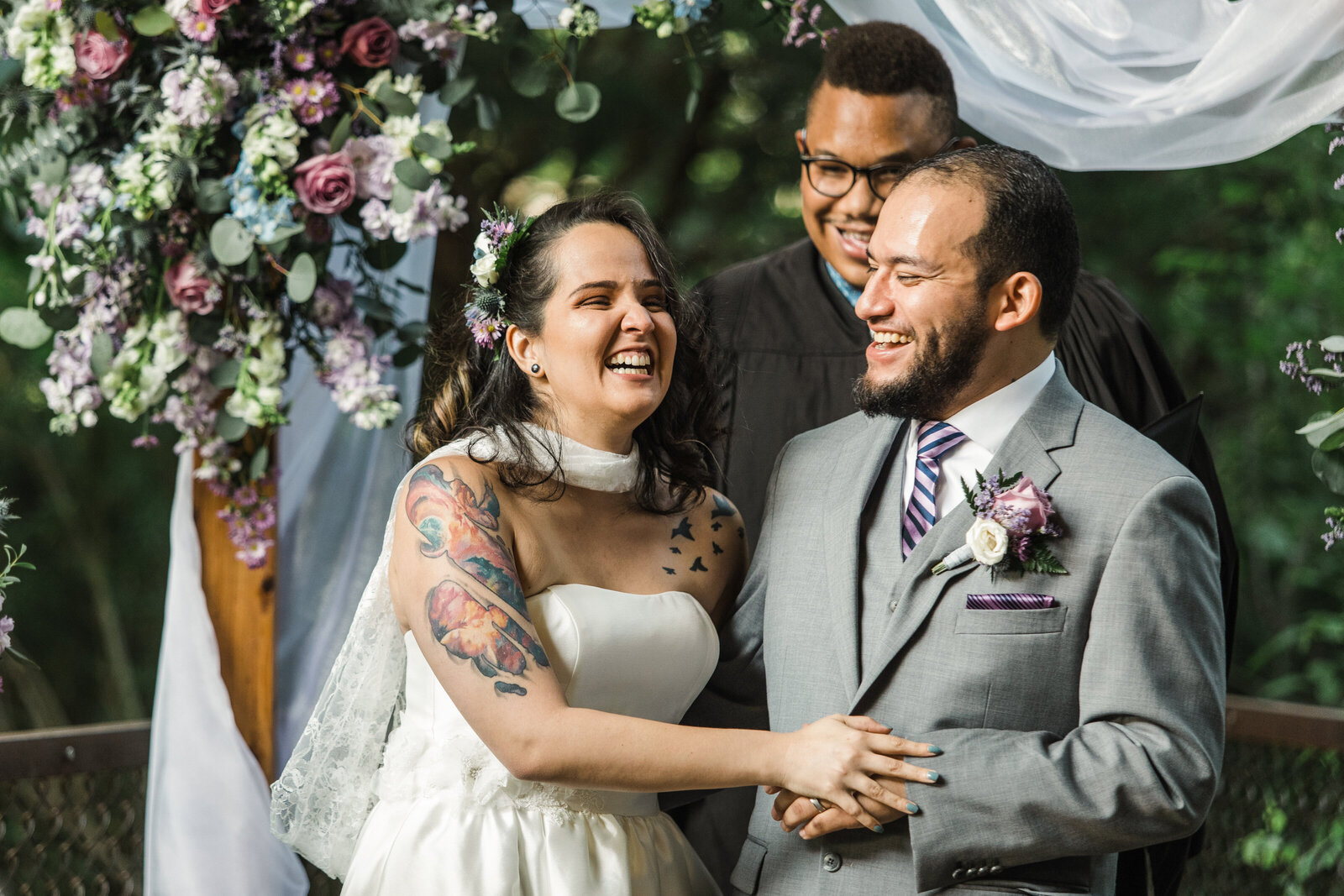 A newlywed couple smiles and laughs after being announced as husband and wife. They are standing in front of an altar with lush purple flowers and white draping. Their officiant is a tall African American man with glasses, and he's smiling. The bride is on the left wearing a bridal cape around her neck that goes down her back. She has a tattoo of a galaxy on her right arm, and is wearing a strapless dress. Her hand is touching the groom's hand. The groom is on the right side and he's wearing a grey suit with a purple tie and boutonniere.