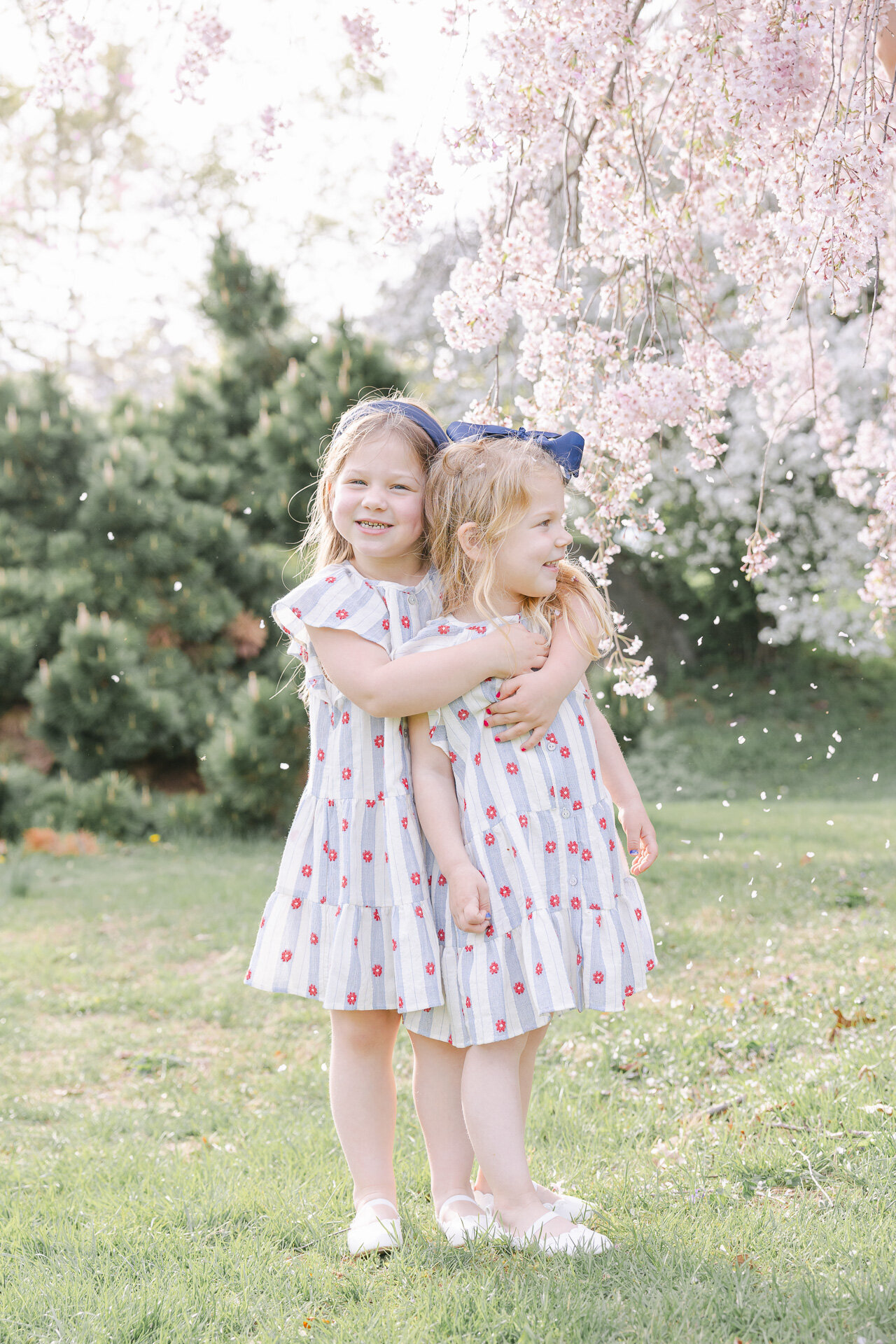 Two young sisters share a joyful hug beneath blooming cherry blossom trees, dressed in matching blue and red sundresses with bows in their hair. Petals float through the air as they smile and play in the spring sunshine, capturing the lighthearted magic of childhood. A perfect example of the playful, story-filled moments from a Greenwich family session.