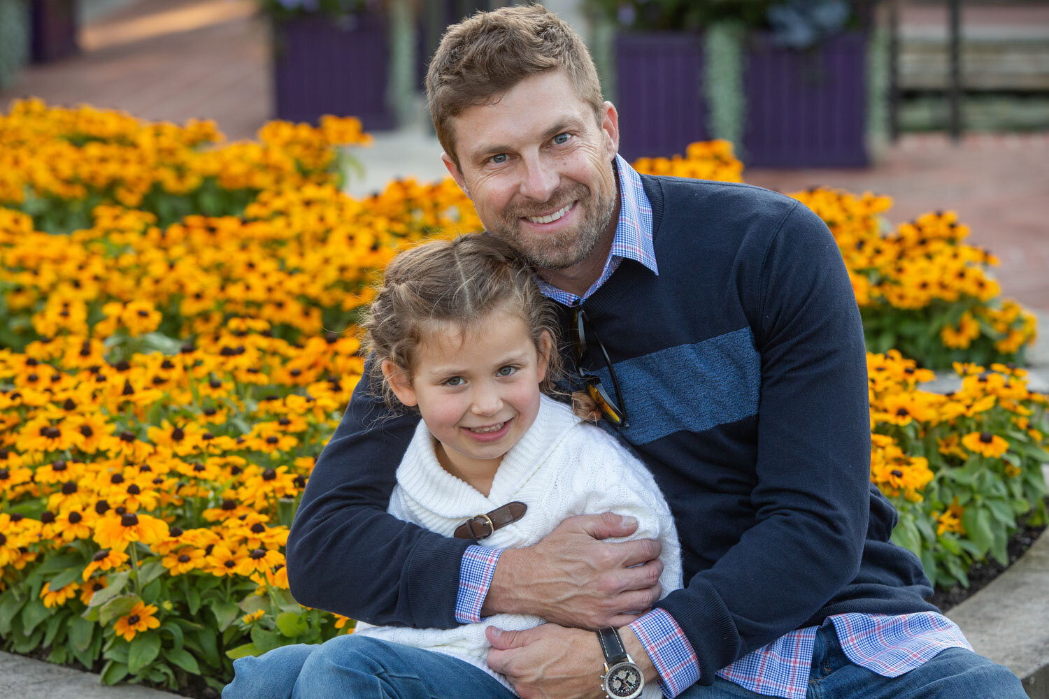 A man is holding a little girl in his arms in front of flowers.