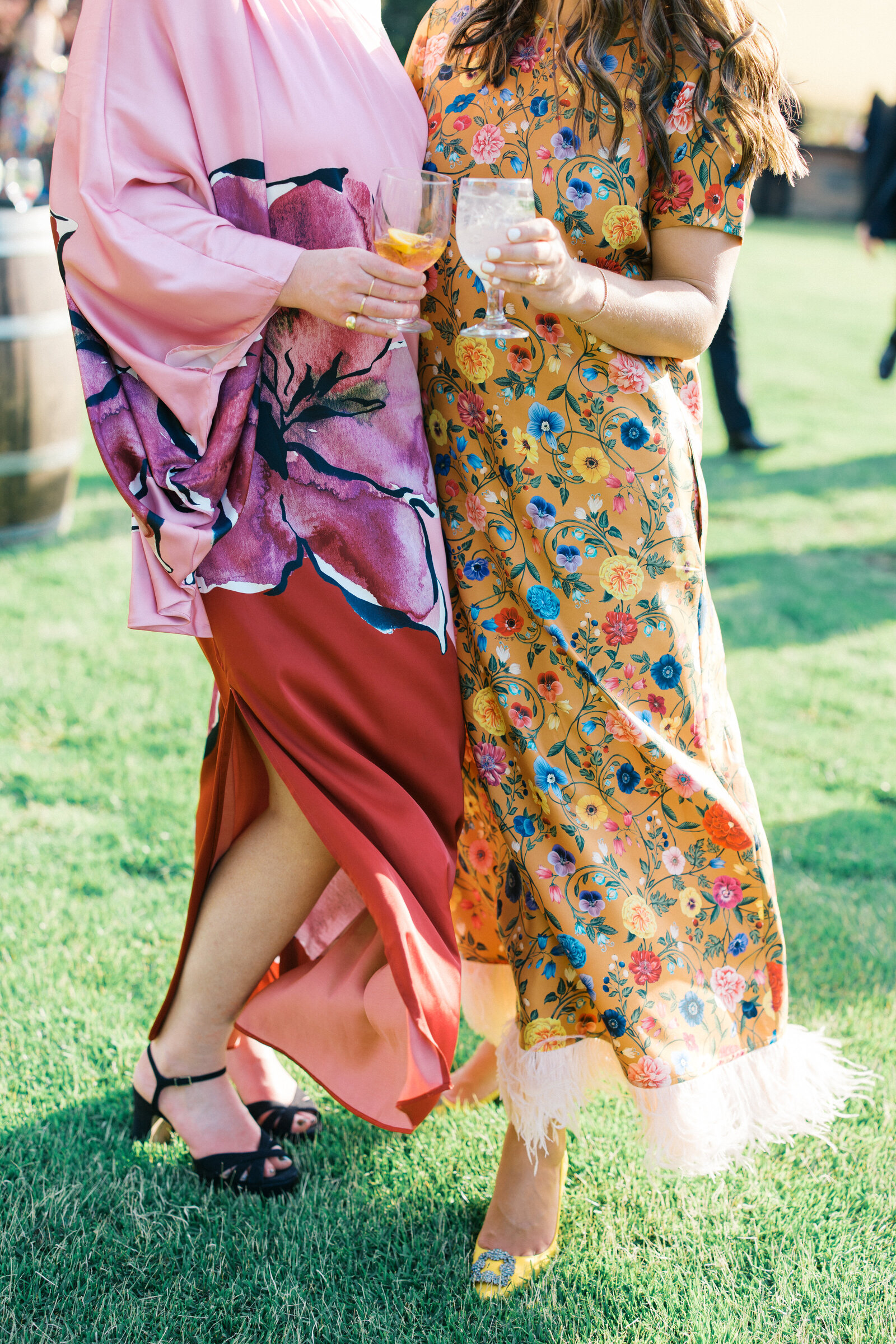 Woman in pink and red dress nd woman in orange floral dress with feather trim and yellow milano feels cheers at wedding cocktail hour on green lawn