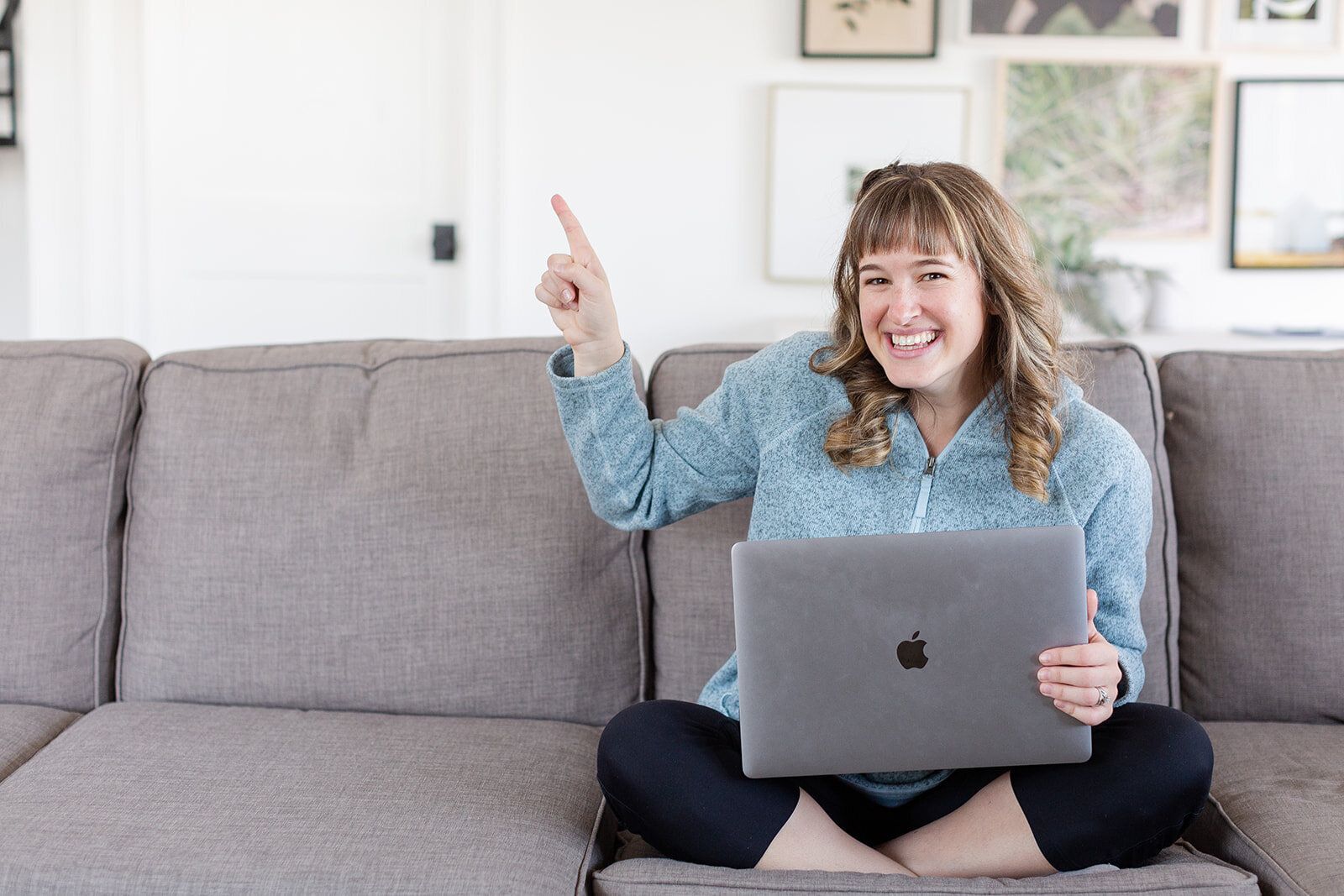 Heather smiling at camera and holding laptop while sitting on couch and pointing her right pointer finger up towards the sky.