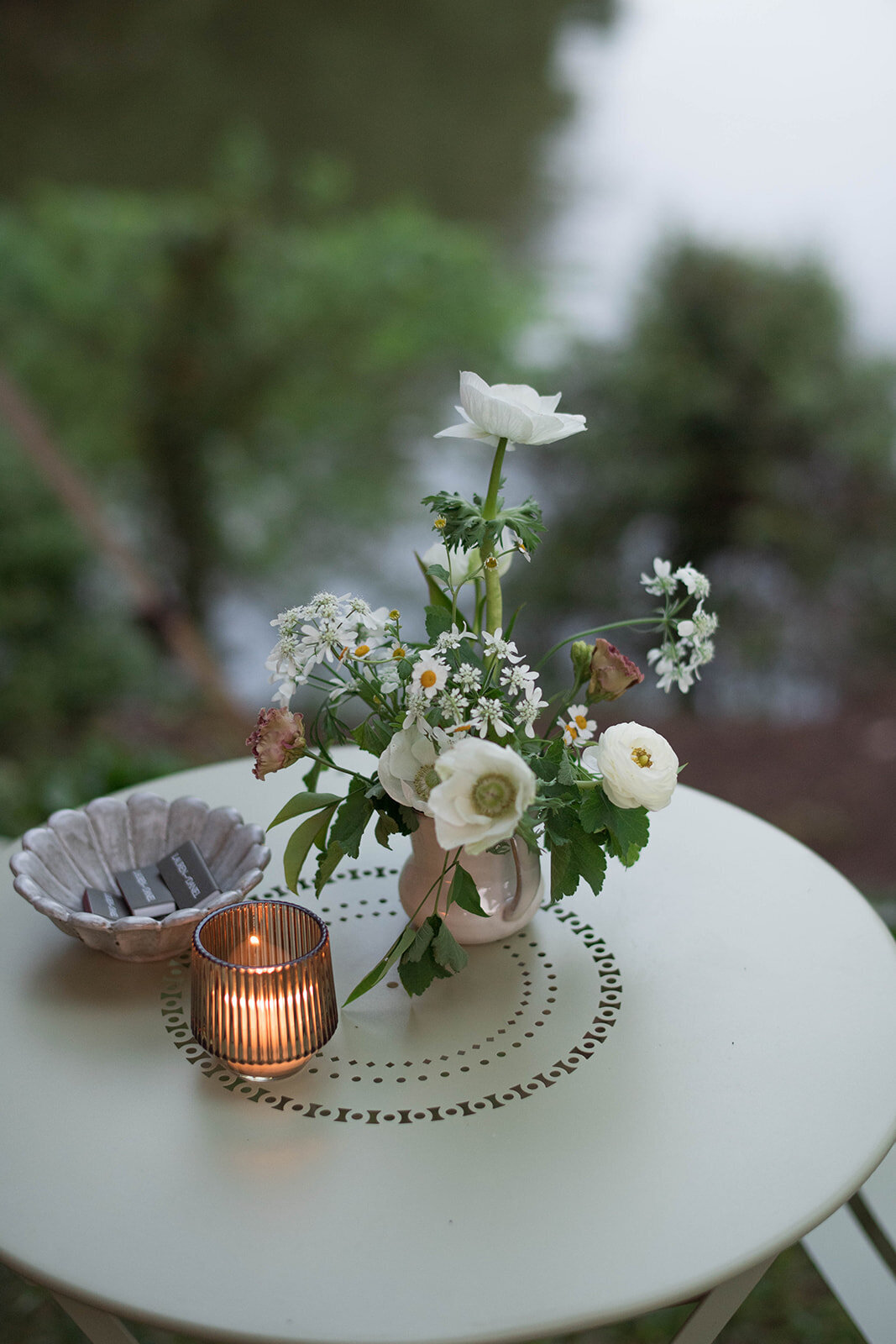 Earthy cocktail table flower arrangement with white anemone, white ranunculus, and daisies. Amber votives and matchbooks on the cocktail table.