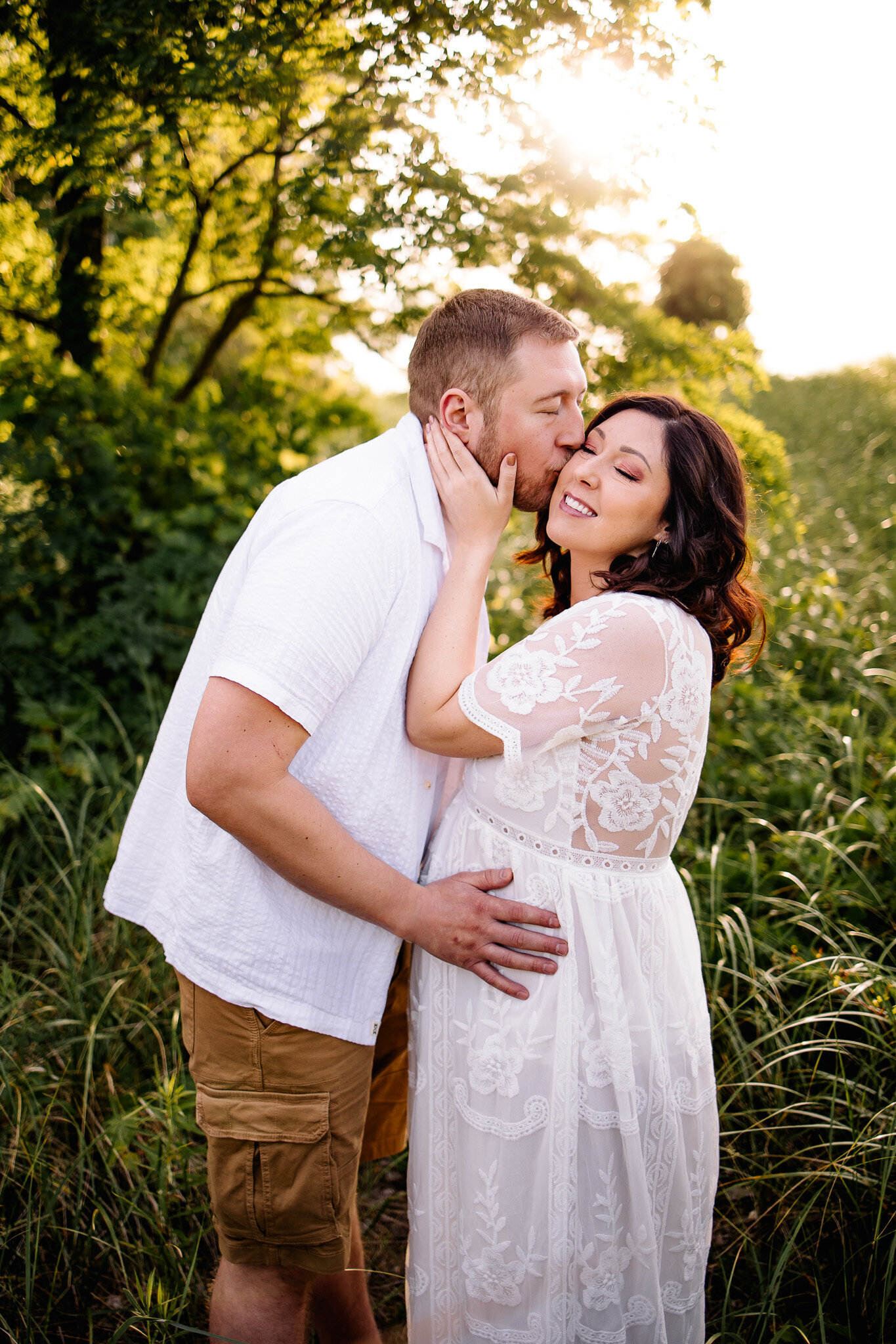 With hand on his wife's pregnant belly, man kisses her on the cheek amongst a field of beach grass