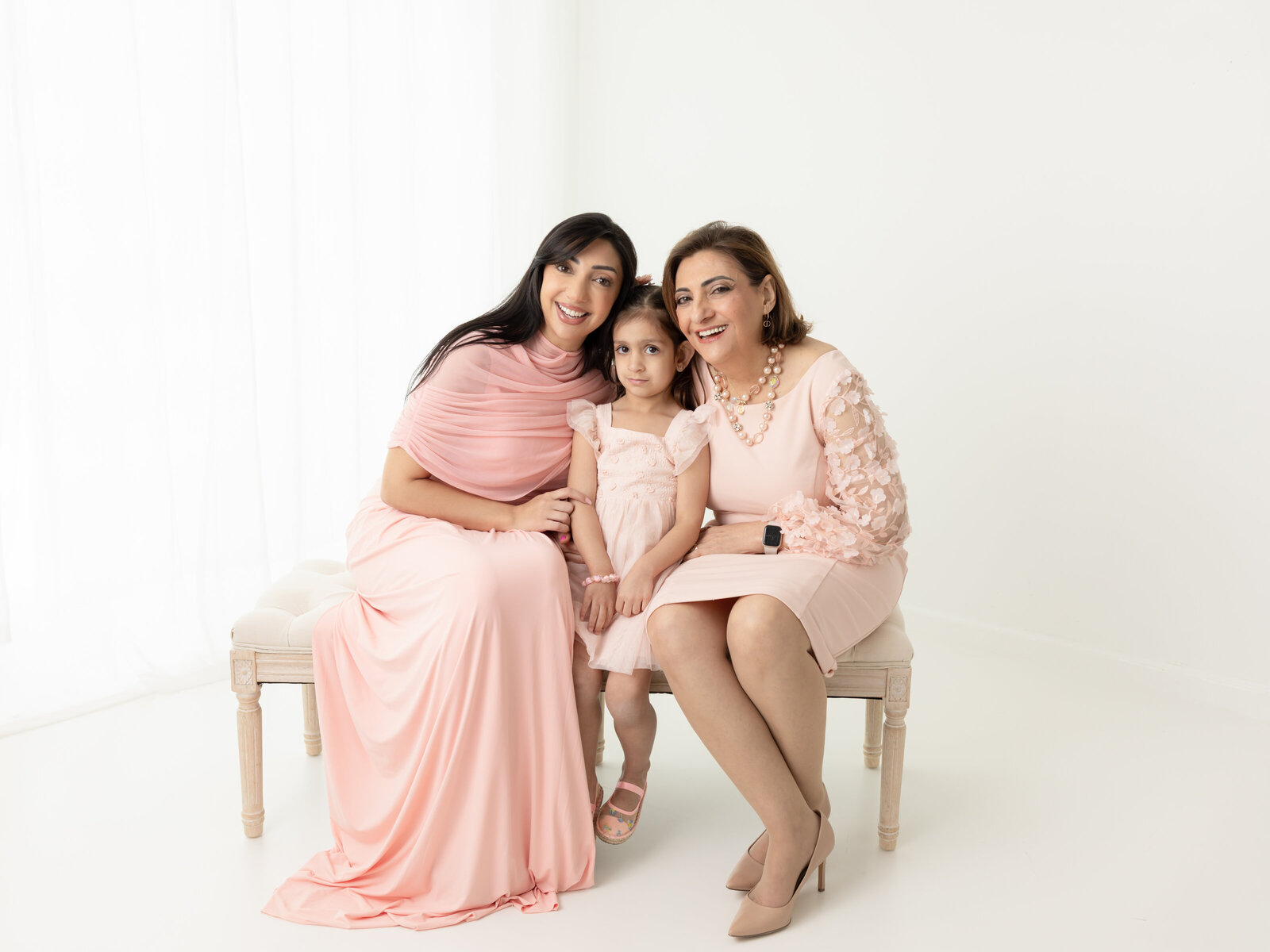 grandmother, daughter and granddaughter in pink dresses sitting on bench for white studio portraits