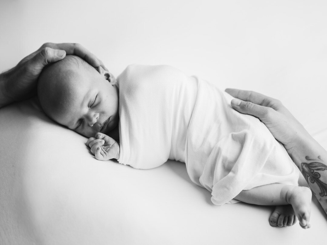 black and white photo of newborn baby wrapped in white swaddle with mom and dads hand on either side of babys body