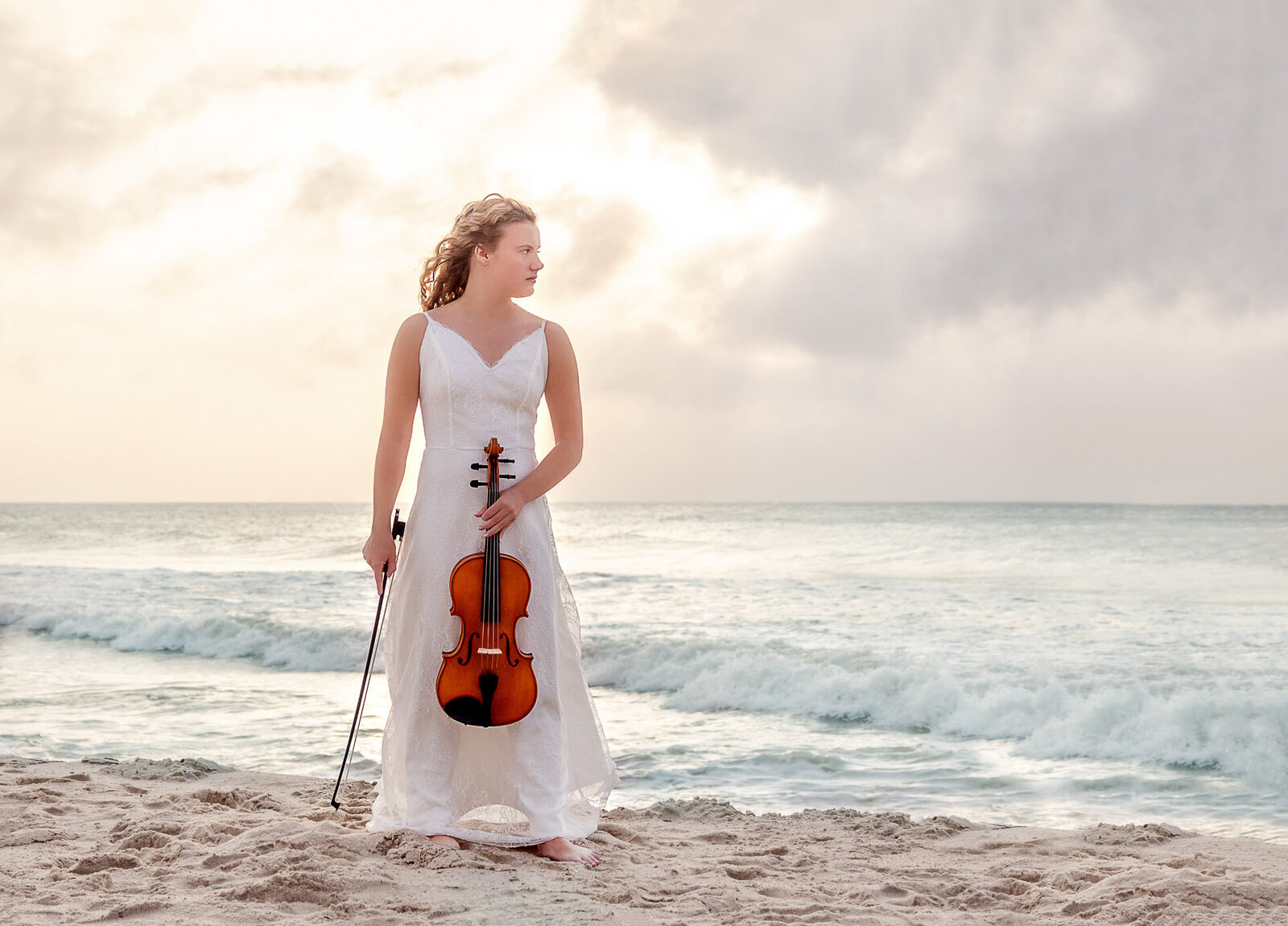 A senior in a white dress holding a viola, standing on the beach with waves in the background in North Carolina. This artistic photo captures the blend of music and nature, ideal for those seeking unique senior photography in Wilmington, NC.