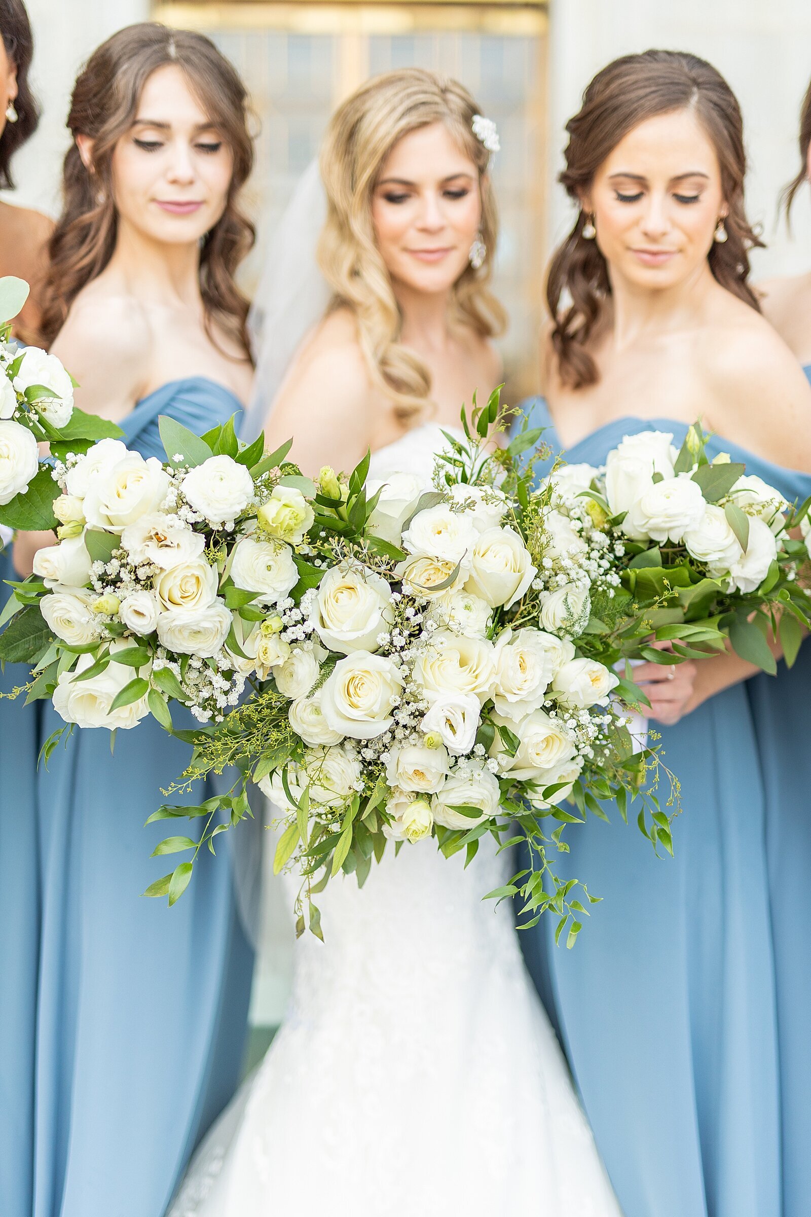 Bride in white lace gown and bridesmaids wearing Azazie blue dresses holding white flower bouquets.
