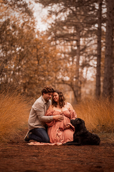 Een aanstaande moeder en haar partner poseren liefdevol met hun hond tijdens een zwangerschapsshoot, een intiem moment vol warmte en verwachting