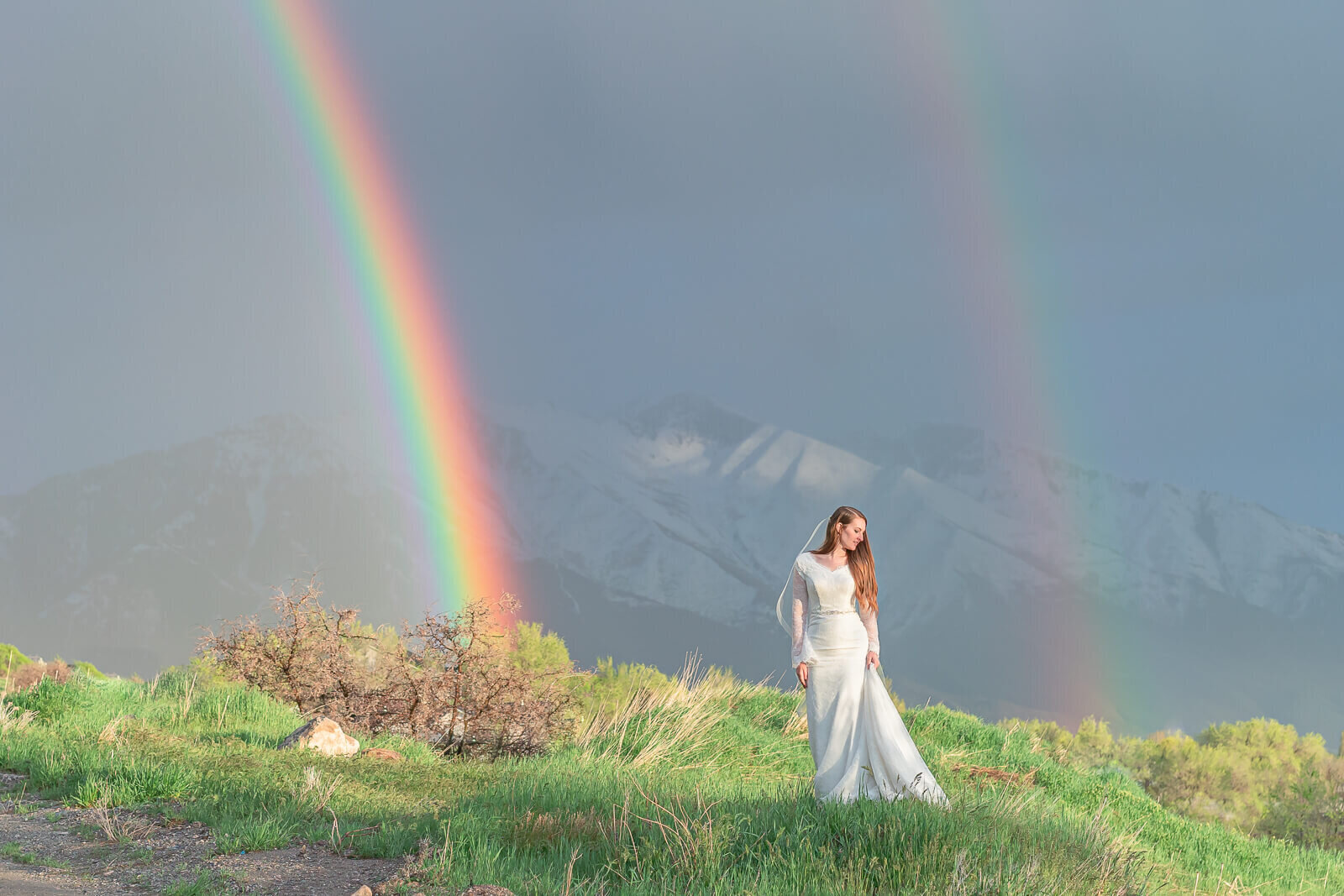 A bride standing in a field of green grass with a double rainbow behind her. Captured near the border of Provo and Springville
