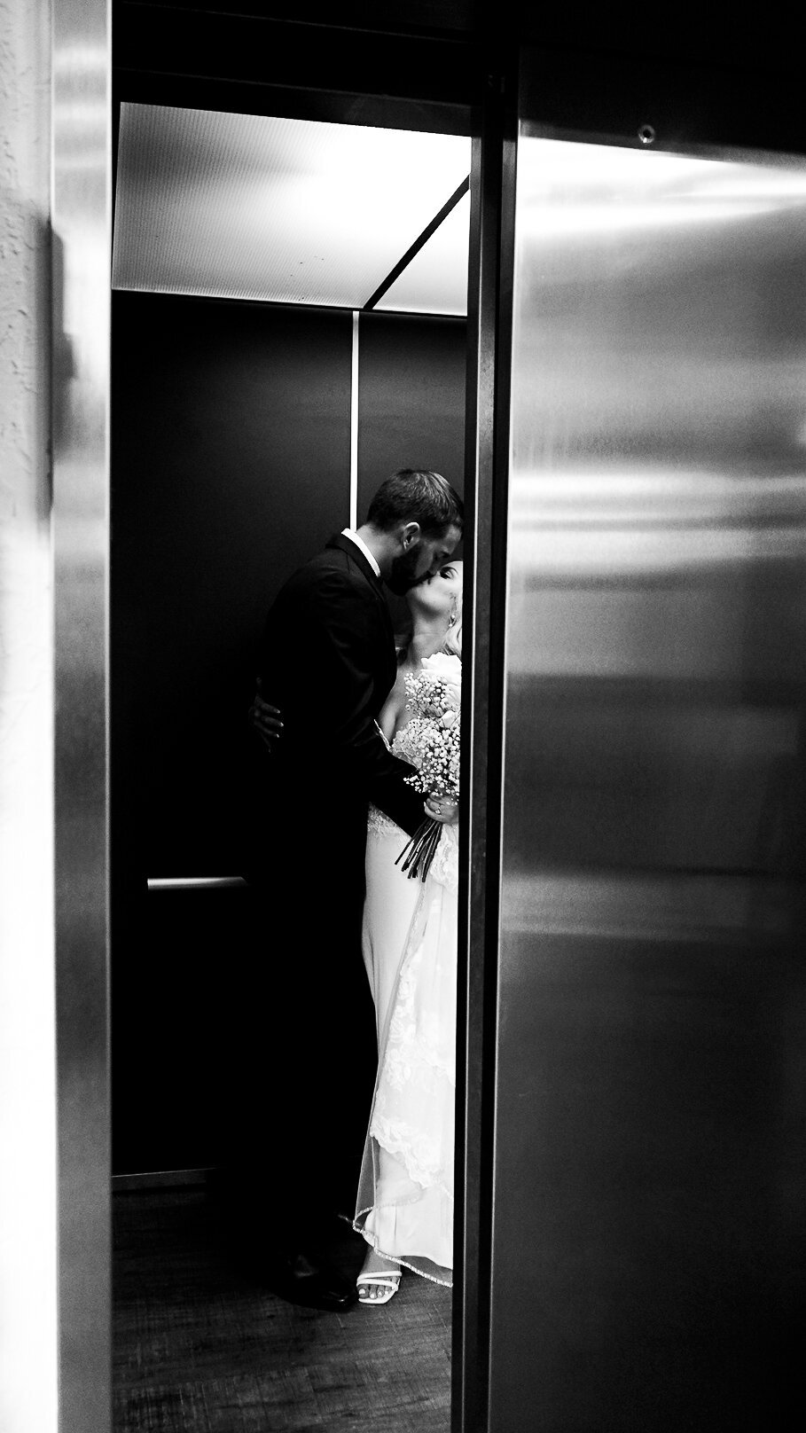 Bride and groom in the elevator at Silver Oaks Chateau, kissing. She is holding a babies breathe and white rose bouquet. The doors are almost closed.