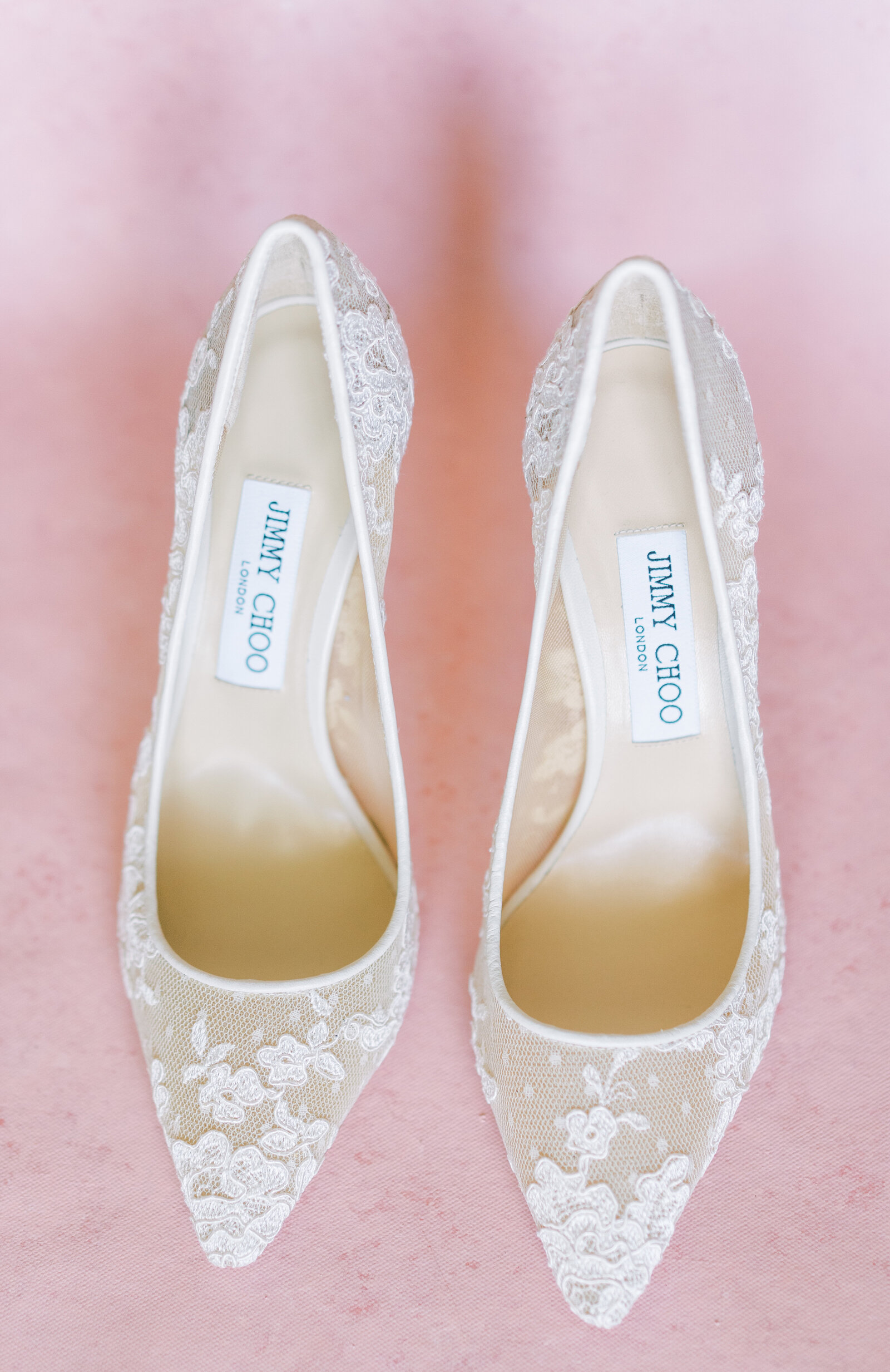 Portrait of white lace Jimmy Choo wedding slippers atop a pink surface