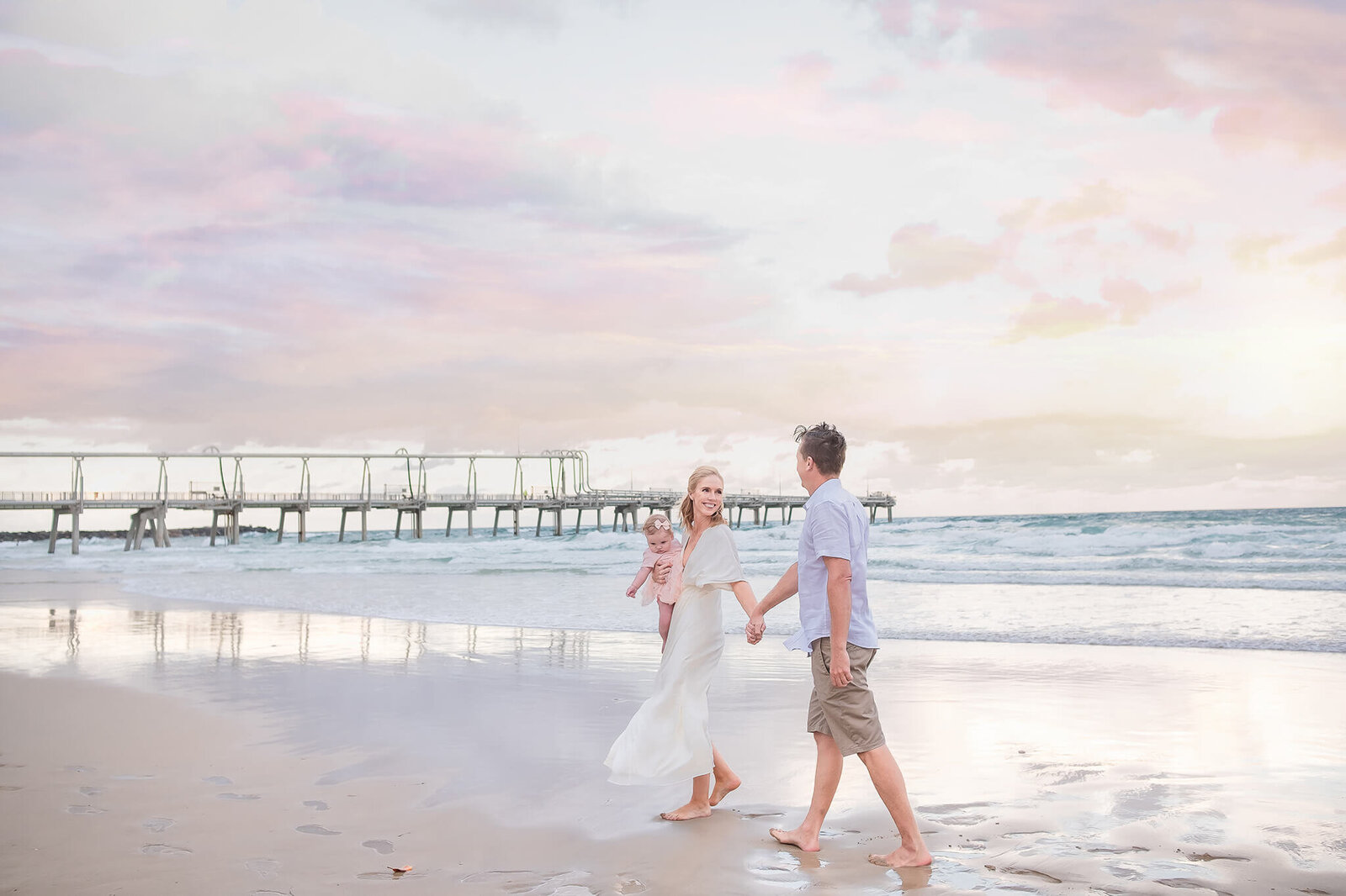 family of 3 walking on the beach with pastel pink sunset backdrop near brisbane