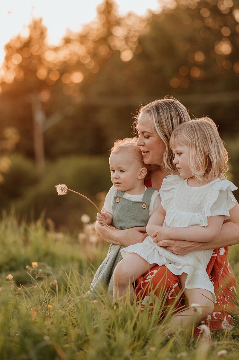 Mom snuggling with toddler son and daughter blowing dandelions in a field. Vancouver Family Photographer