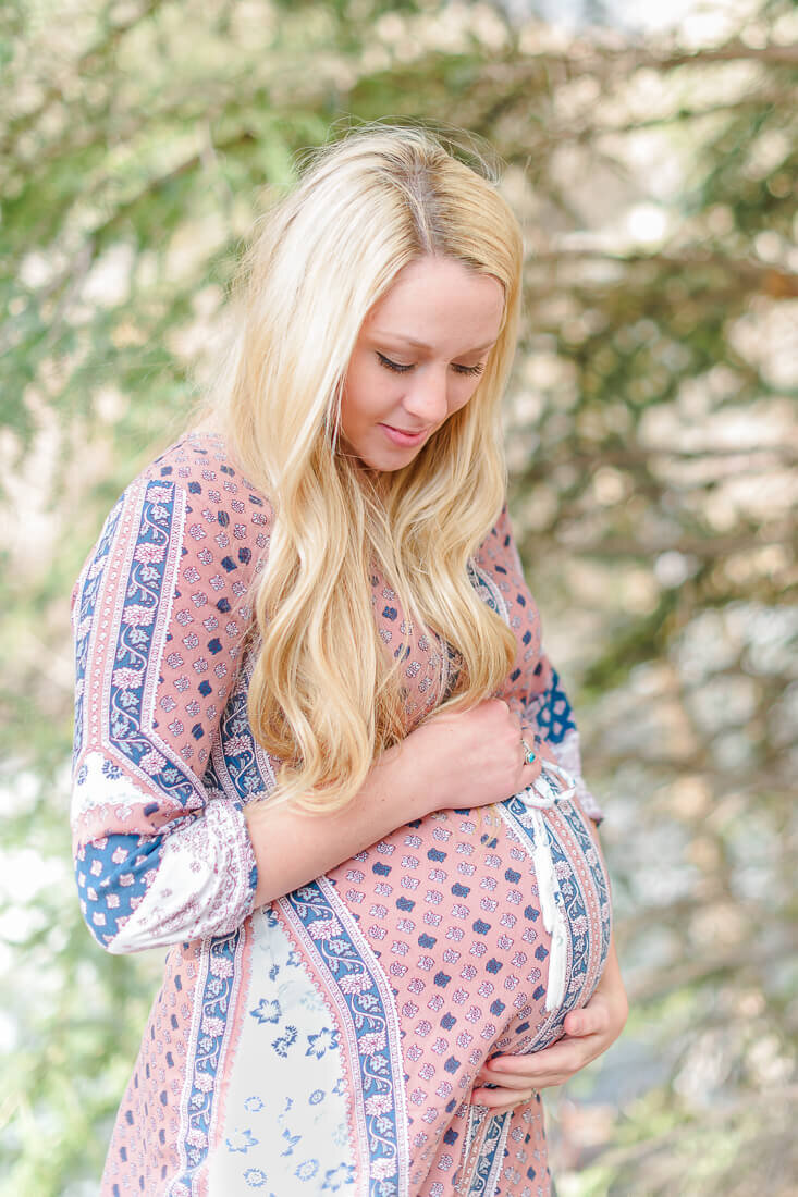 A pregnant woman with long blonde hair wearing a pink maternity dress holds her belly stands in front of pine tree branches in Provo Canyon in the spring
