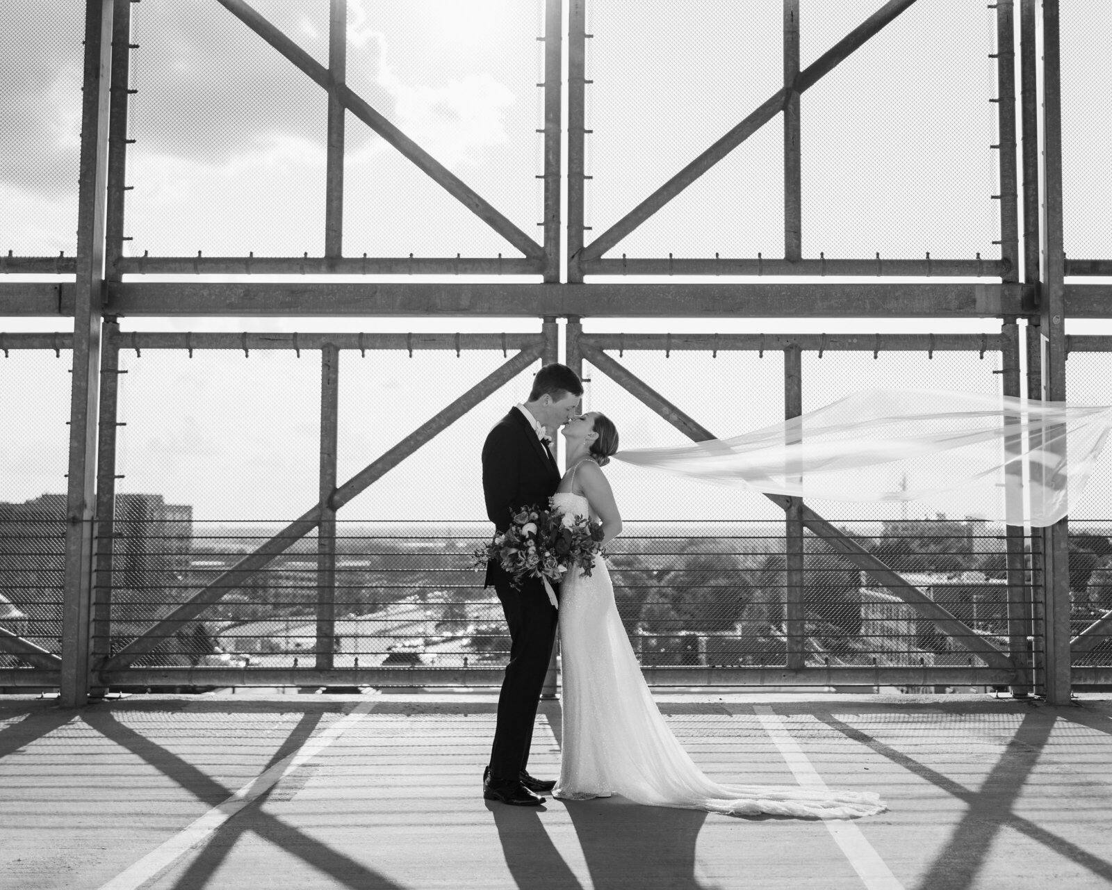 Dramatic black and white photo of a bride and groom sharing a kiss on top of a parking garage while the bride's veil sails in the wind
