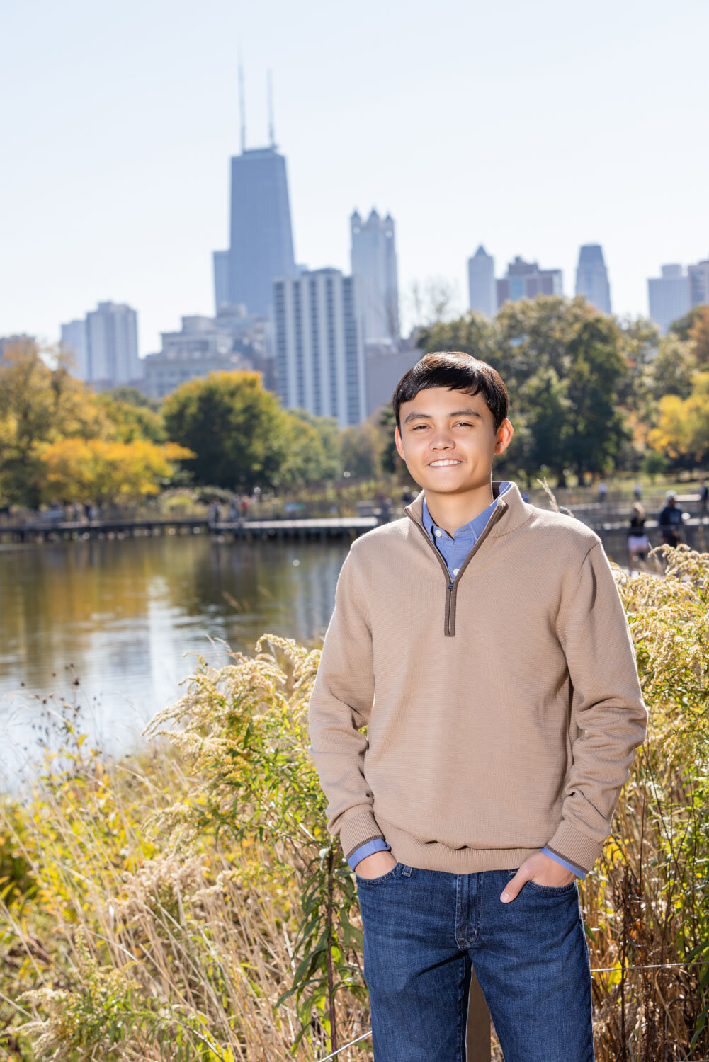 A young man in a tan sweater stands in front of a body of water