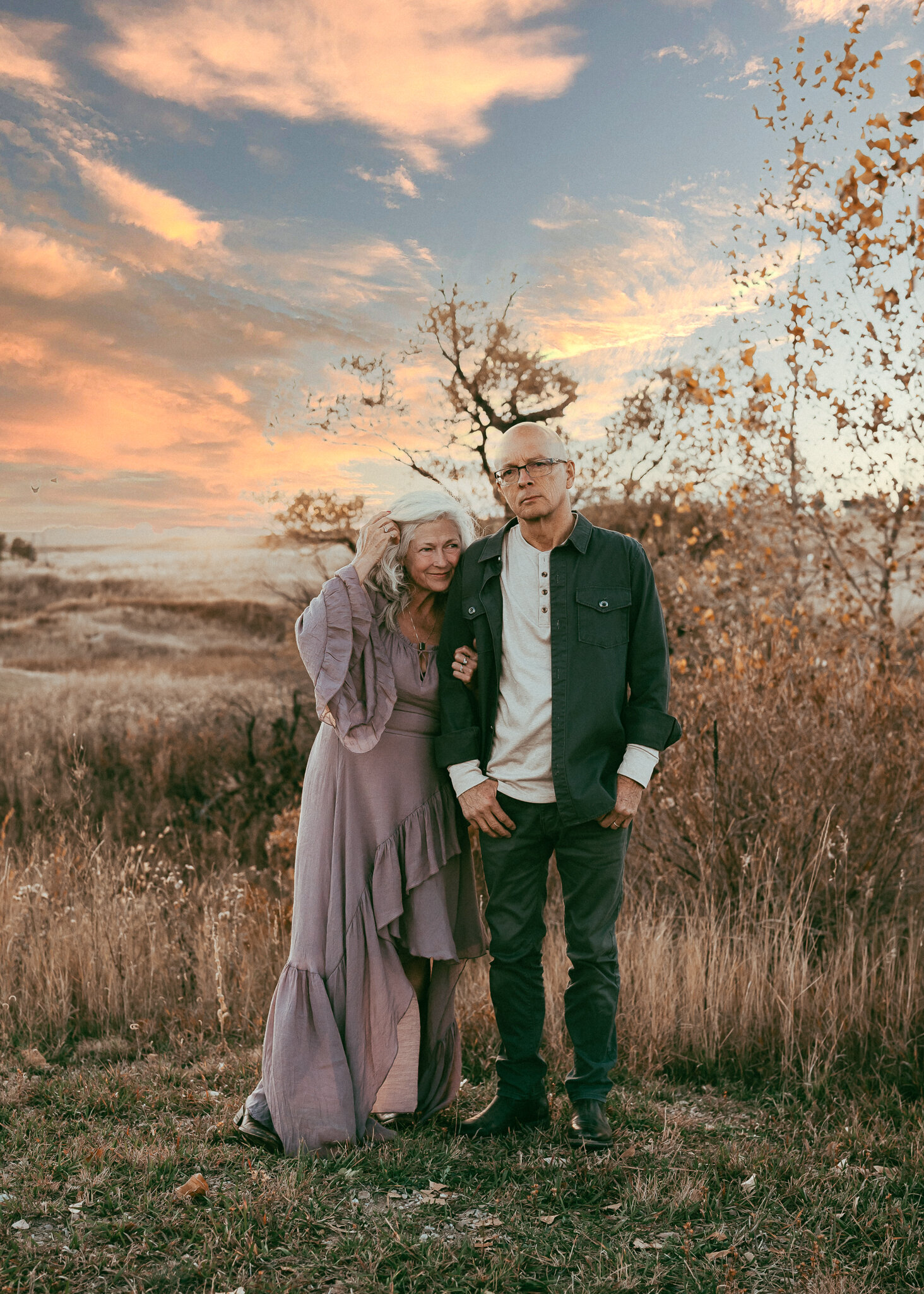 A couple that is grandparent age stands together in a fall mountain setting the wife is wearing a lavender dress while the husband is dressed like an urban cowboy.