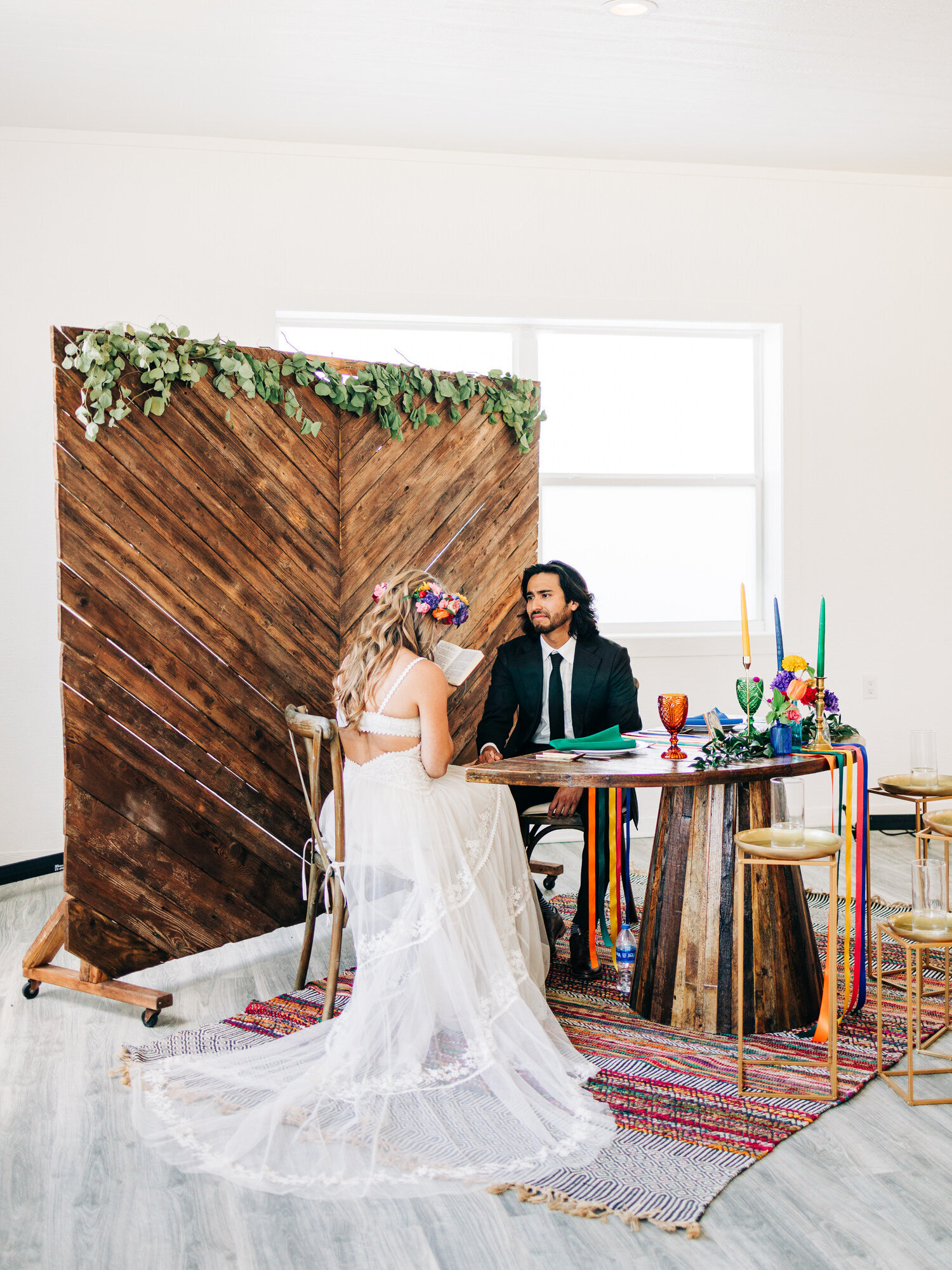 A bride and a groom sit at their sweetheart table in an all-white reception venue. The table is decorated with colorful candles and ribbons. The bride is wearing a flower crown and a boho wedding dress. The bride and groom are reading letters to each other before their wedding ceremony. This image was taken by a wedding photographer in Austin, TX.