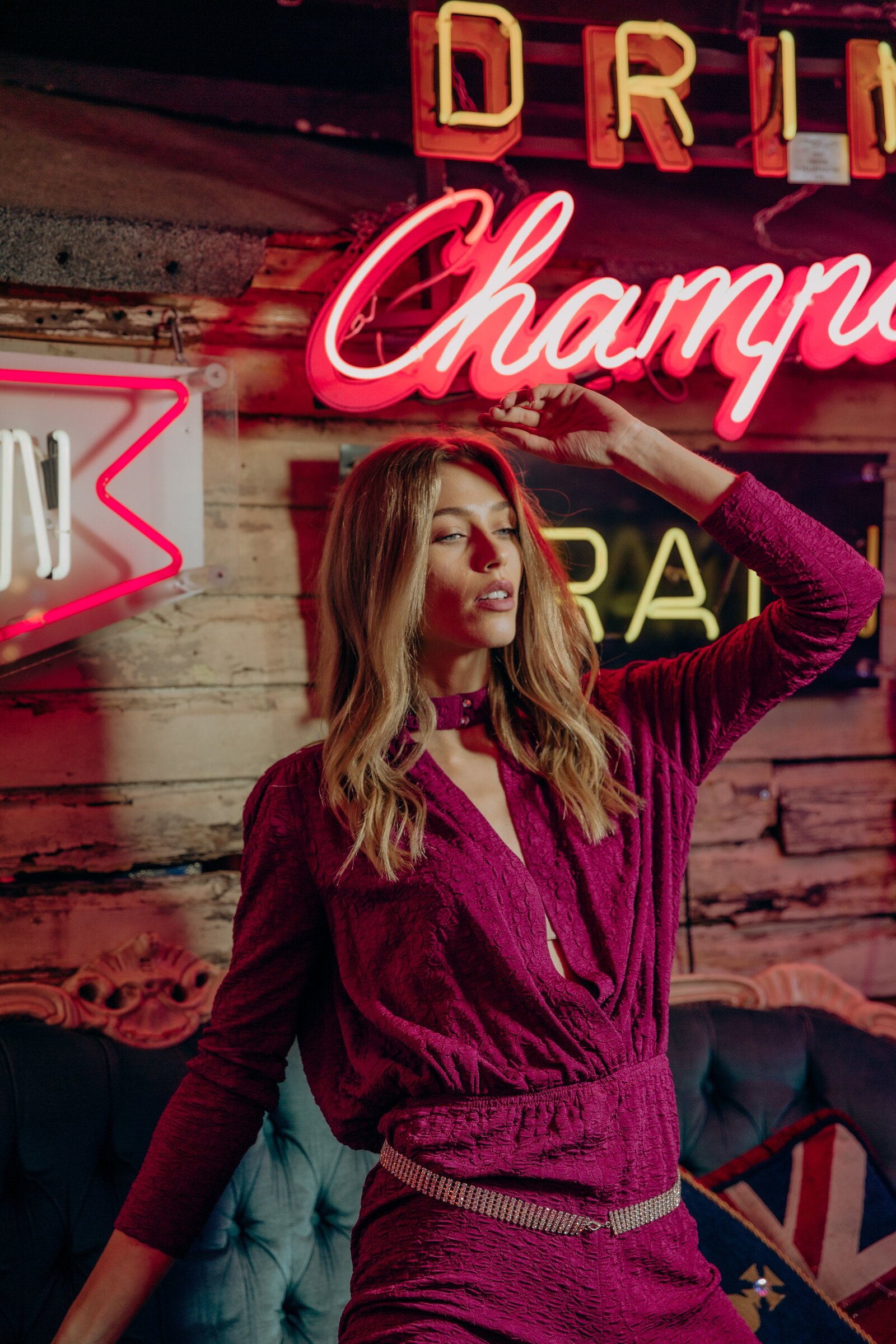 Anastazja wearing a pink jumpsuit kneeling on a blue sofa surrounded by neon signs.