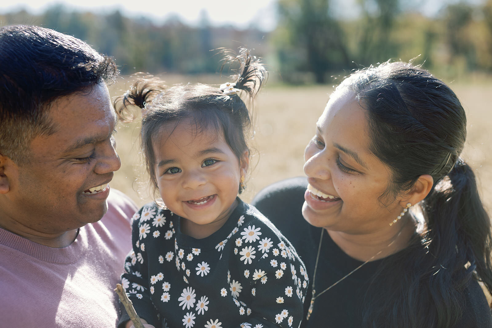 Playful outdoor family portrait with a laughing toddler in pigtails surrounded by smiling parents under soft sunlight.