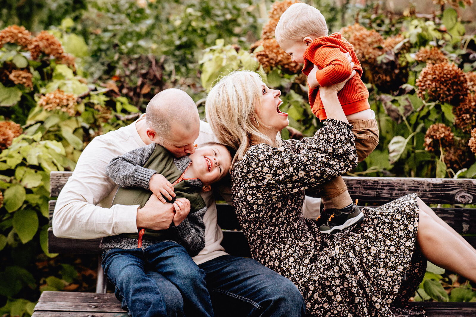 A lively family moment captured on a park bench, where a mother playfully lifts her toddler in the air while the father kisses their older child.