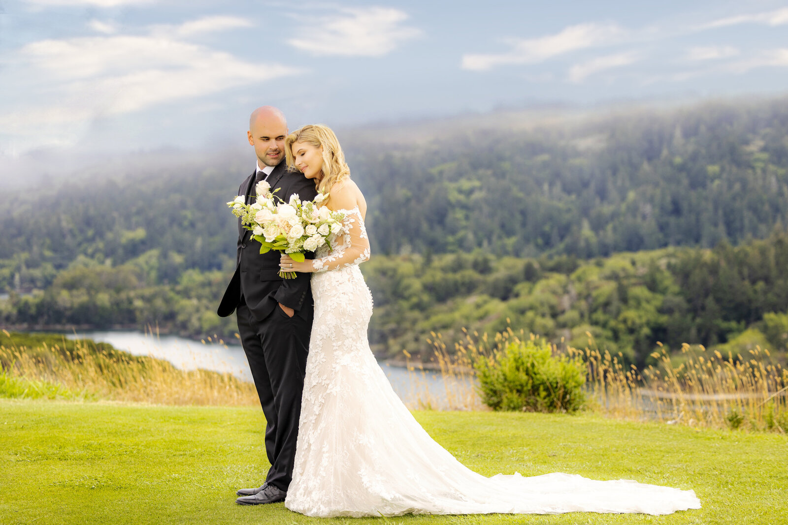 A bride hugs groom from behind in front of green mountains and lake. Photo by Sacramento photography studio, philippe studio pro.