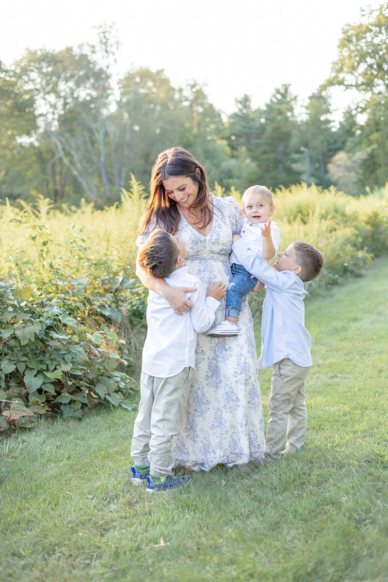 Mother stands in a wildflower field lovingly surrounded by her three young sons, holding her toddler and smiling down at the older boys as they hug her legs.