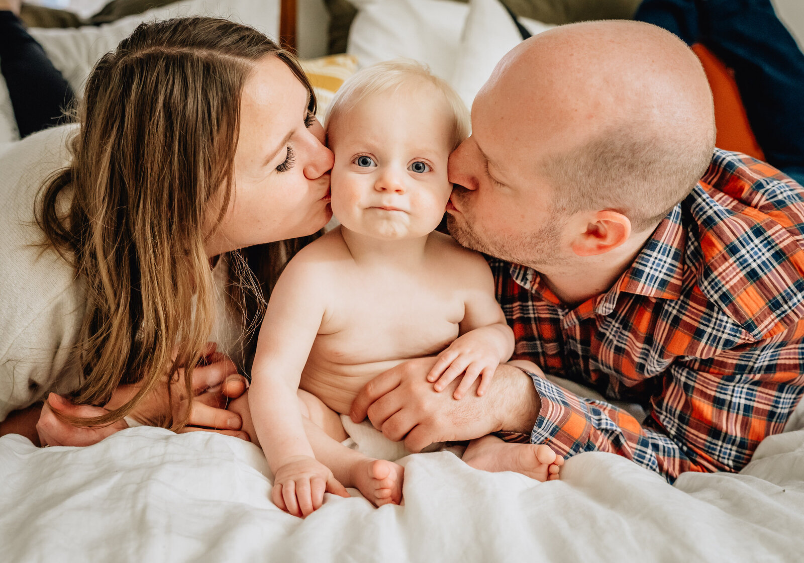A sweet moment where two parents are lying on a bed, each kissing the cheeks of their smiling baby boy, who is sitting between them.