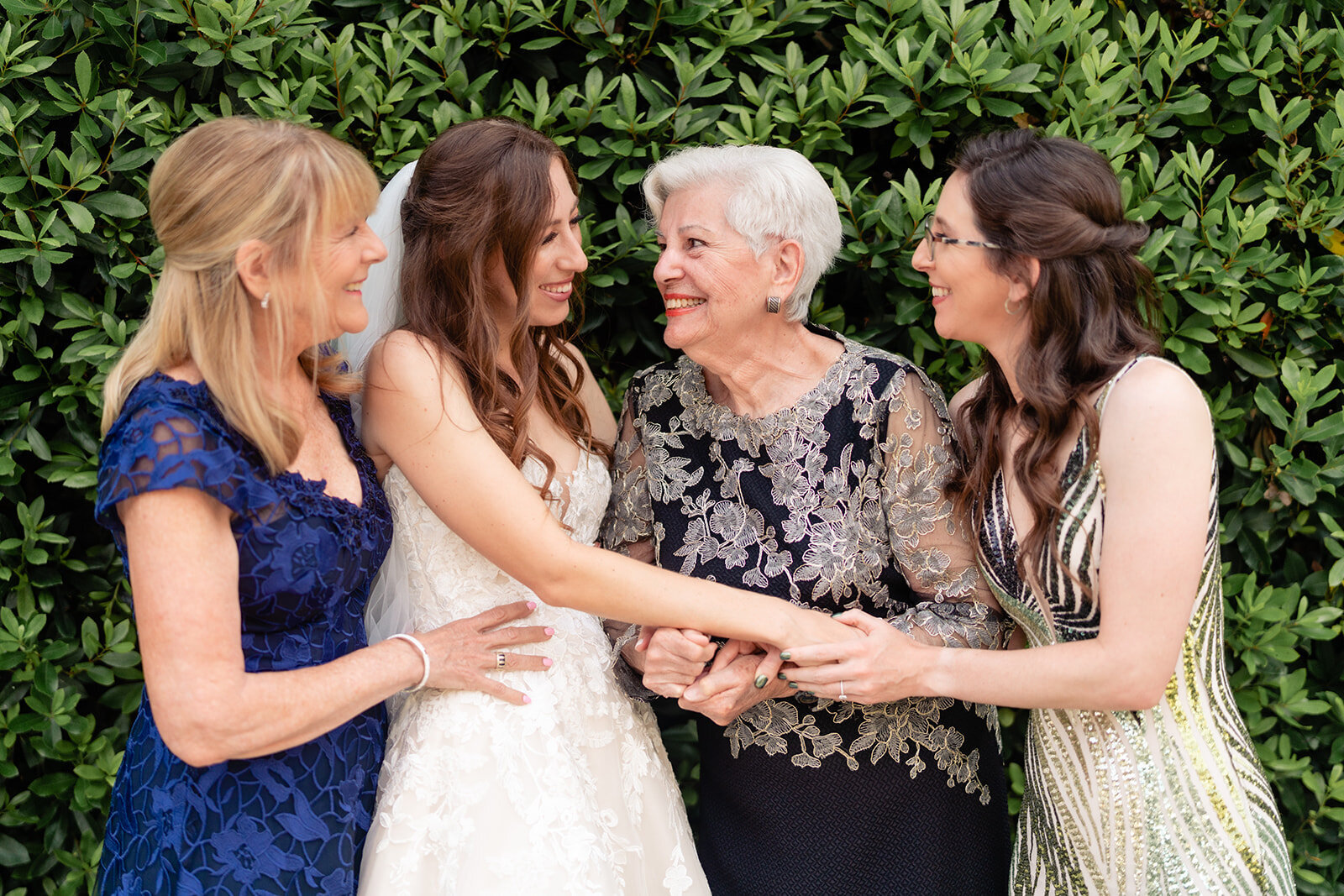 A bride being embraced by her mother, grandmother, and sister in front of a tall green hedge at Estancia