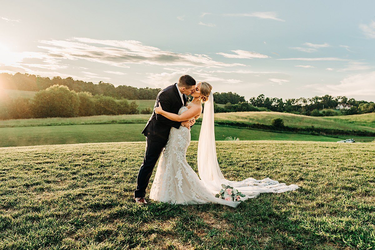 The bride, wearing a long white gown and veil, kisses the groom wearing a navy suit on a hilltop in Tennessee