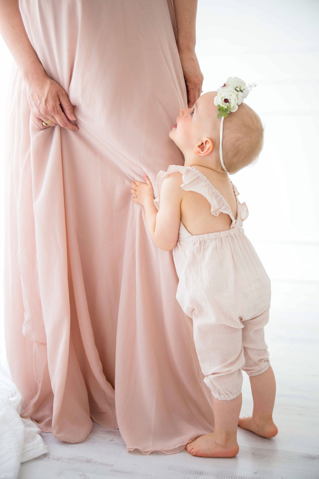 Babyshooting Bielefeld Fotostudio. Kleines Mädchen mit Blumenkranz hält sich an Mamas Rock fest und schaut zu ihr lächelnd rauf.