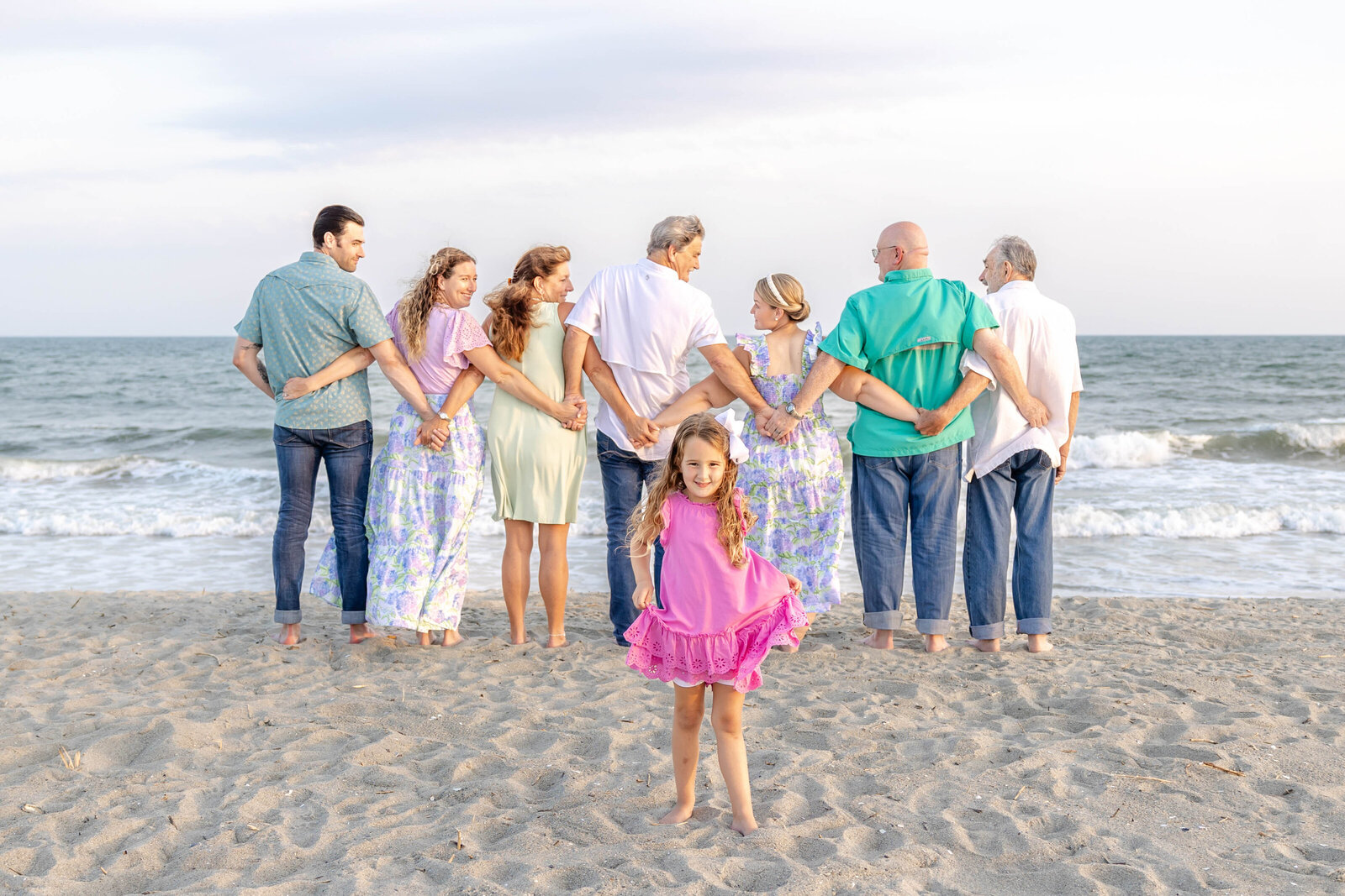 A family stands arm in arm, facing the ocean, while a young girl in a pink dress playfully poses in the foreground. The scene captures a lively beach moment, perfect for artistic family photography in Wilmington, NC, showcasing dynamic family interactions.