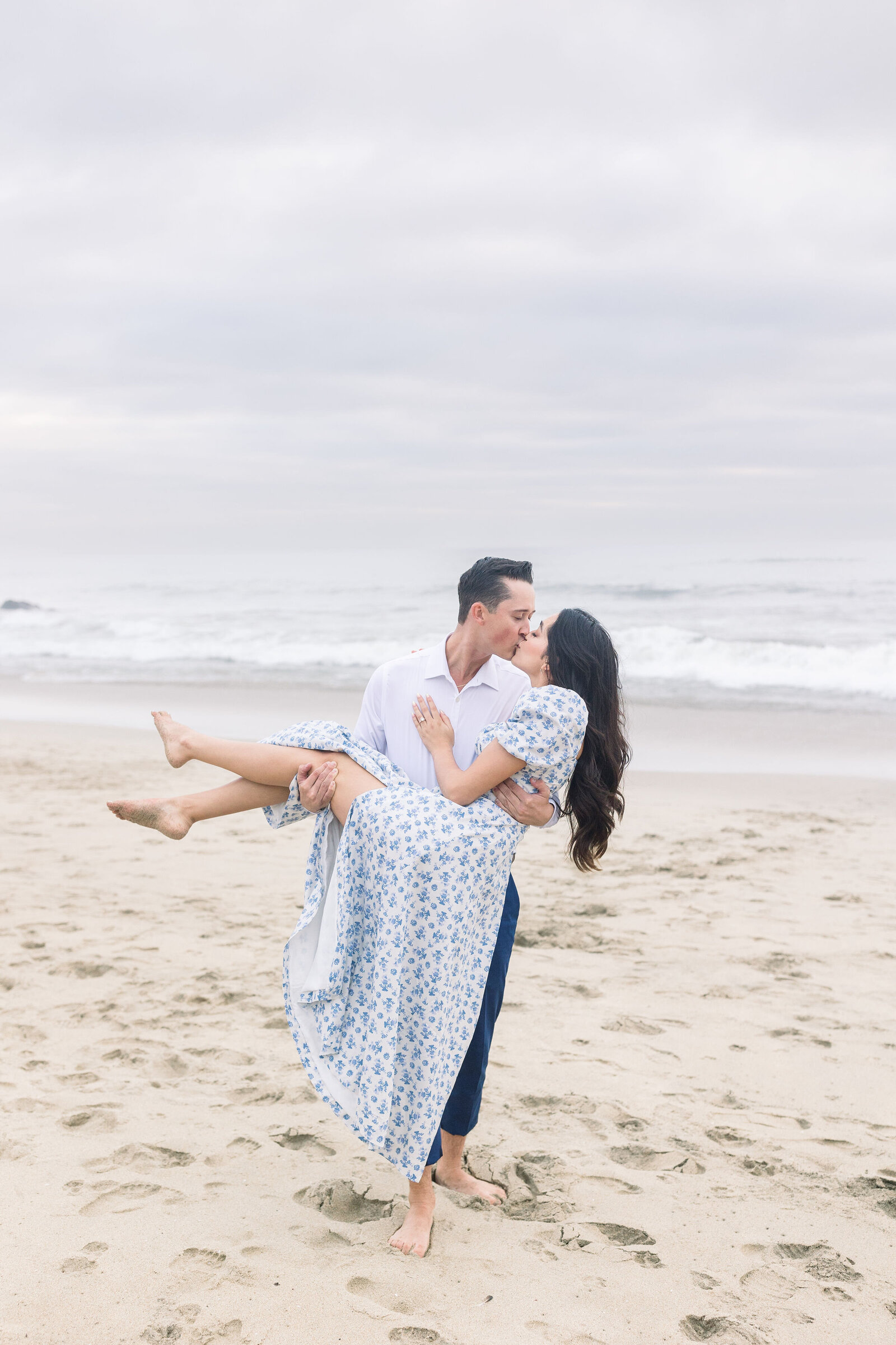 wedding photographer bay area photographs groom carrying a woman while at half moon bay for beach engagement photos with the shore right behind the couple