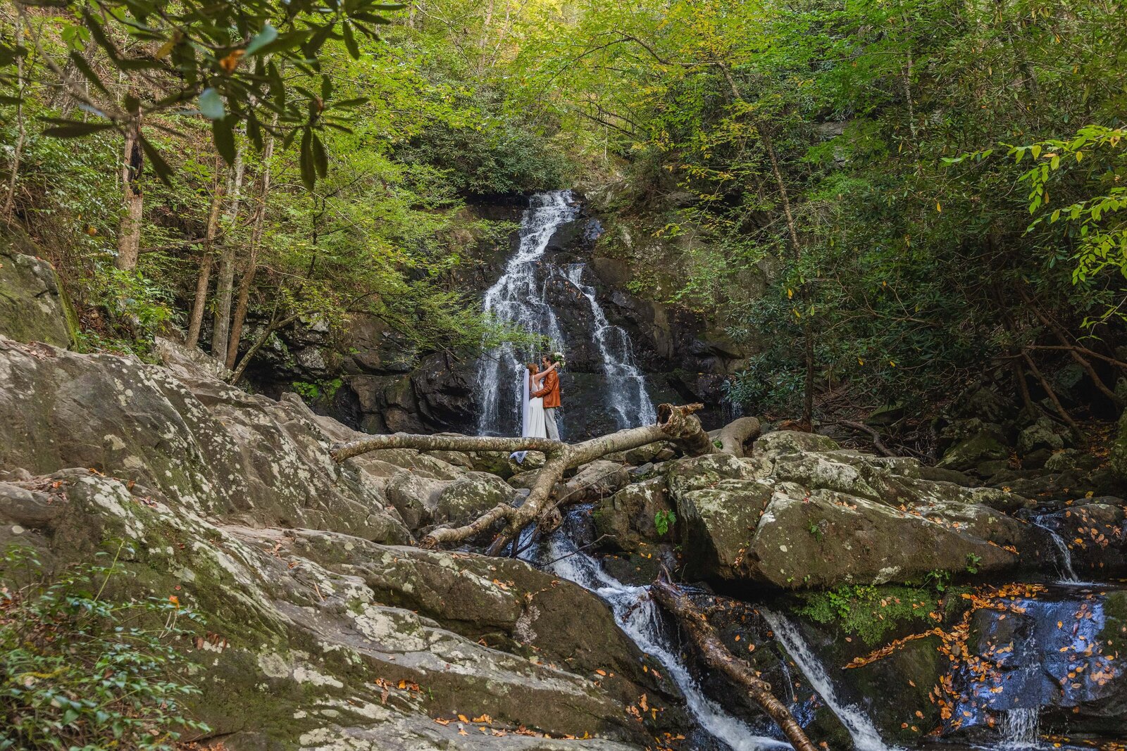 A bride and groom stand in front of a large, tumbling waterfalls sharing a kiss. The bride is in a flowy wedding dress with a long veil and the groom is wearing tan pants with an orange leather jacket. They are surrounded by rocks and lush green trees.