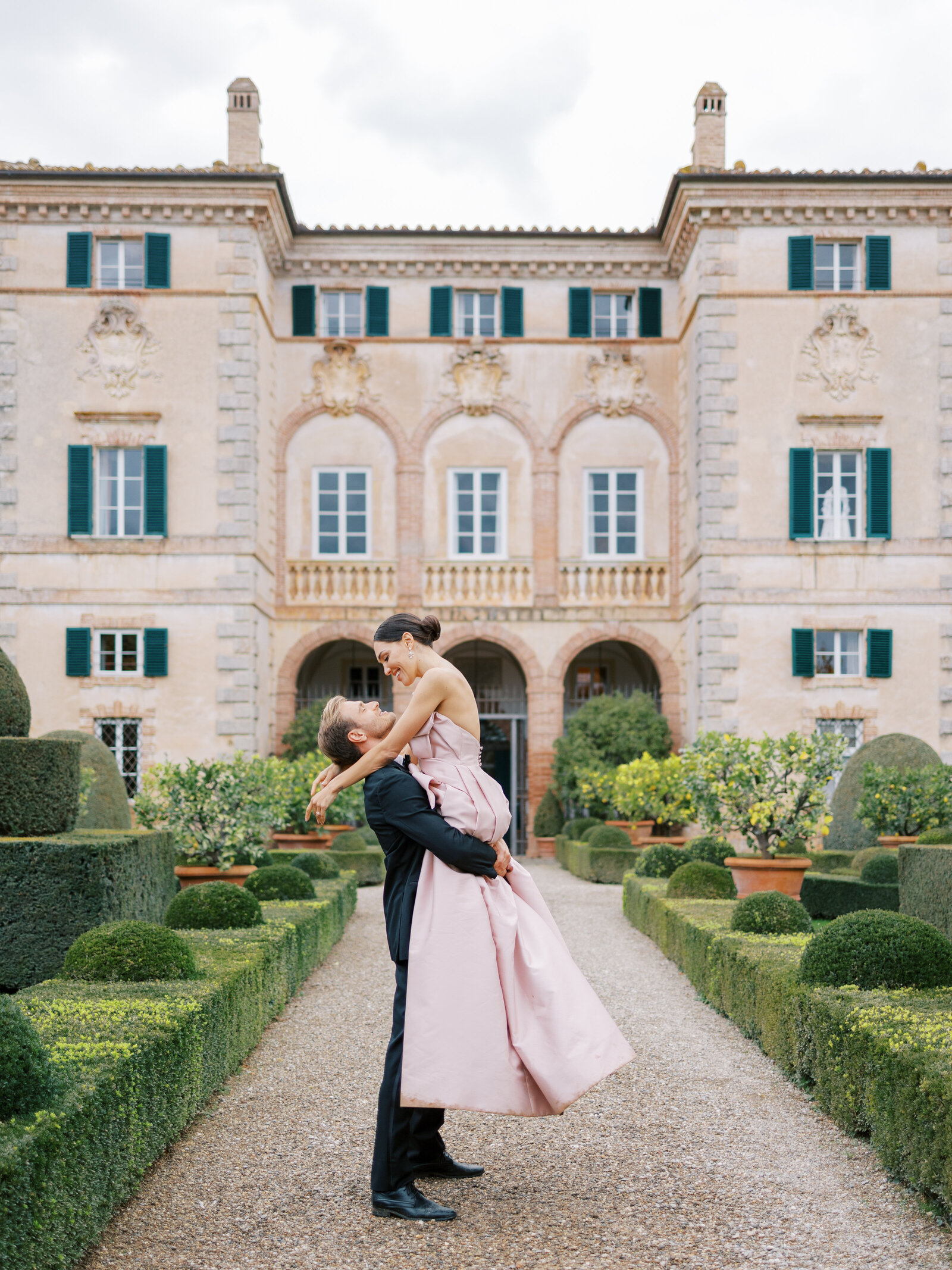groom lifts bride in front of Villa Cetinale in black tux and pink gown on wedding day in Italy