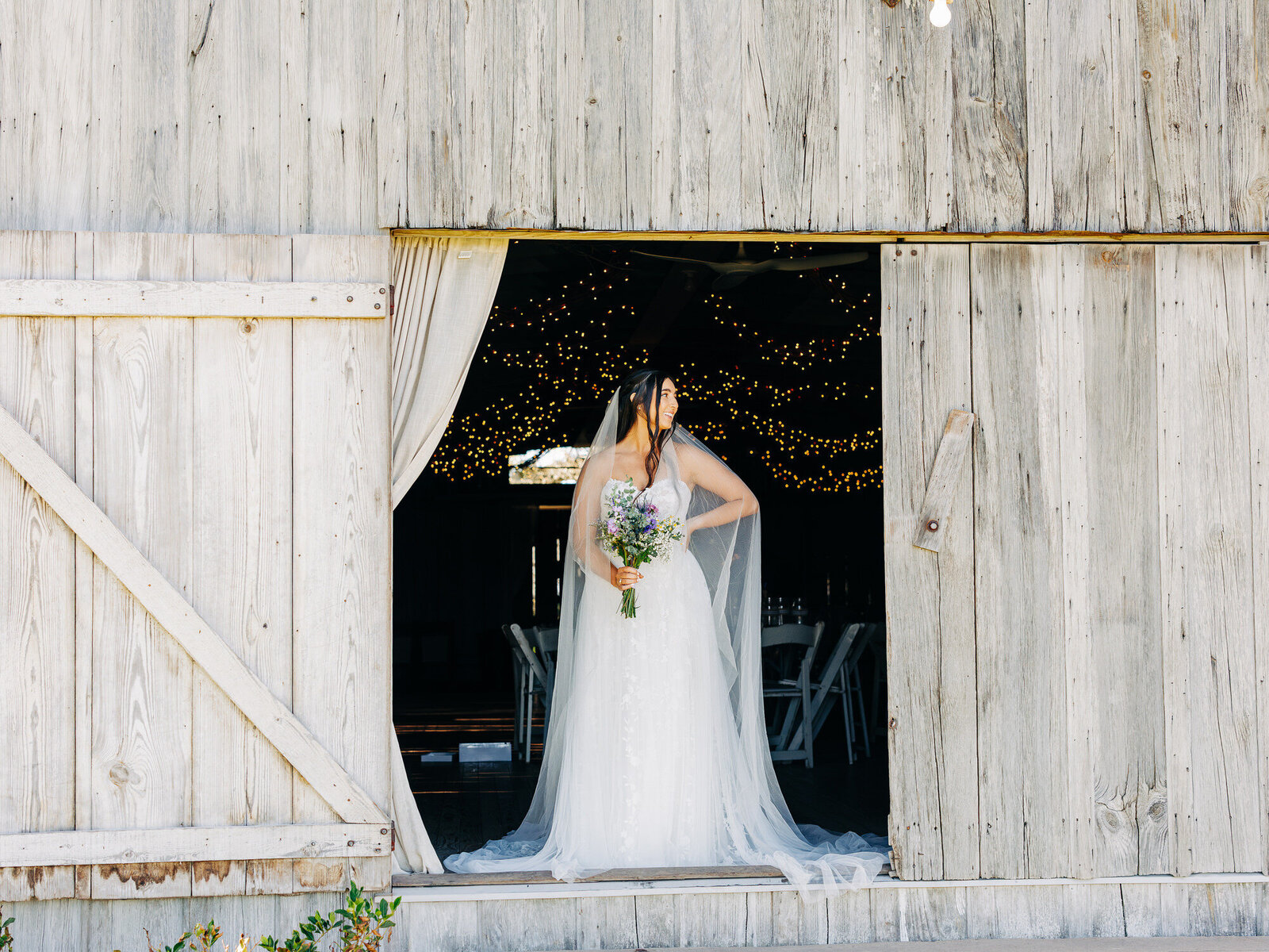 A bride in a white dress and veil stands in a rustic wooden barn doorway, holding a bouquet of flowers. Warm string lights and white chairs glow behind her—captured beautifully by an Eau Claire Wedding Photographer.