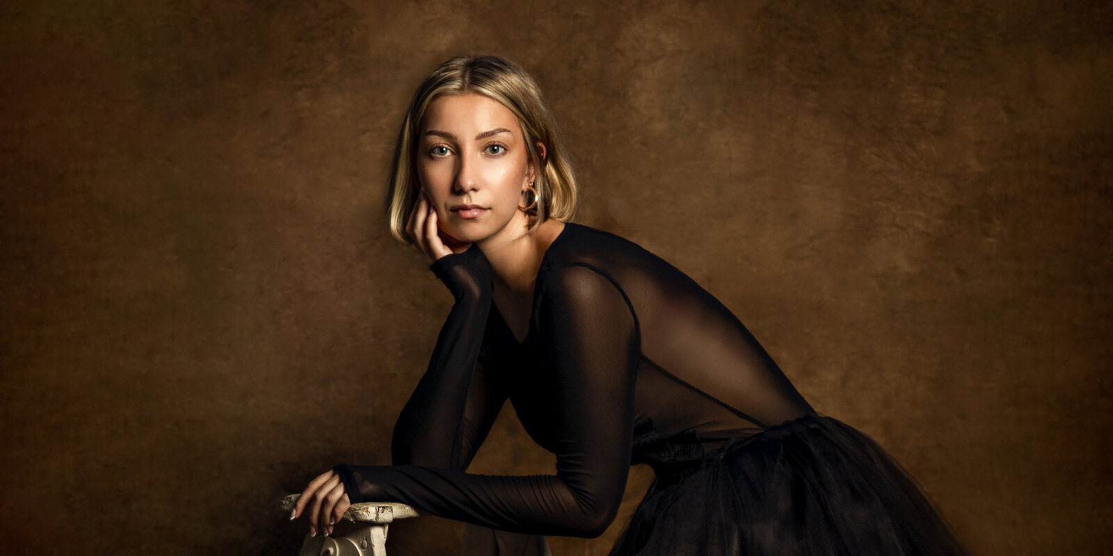 woman in black sheer top leaning on a ladder on custom brown backdrop